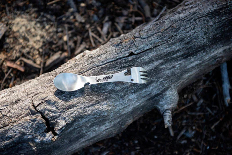 Metal camping spork utensil on weathered log, outdoor adventure gear