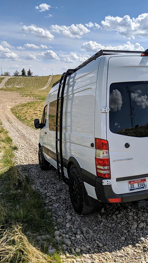Sprinter van with roof rack and ladder on rocky off-road trail under blue sky