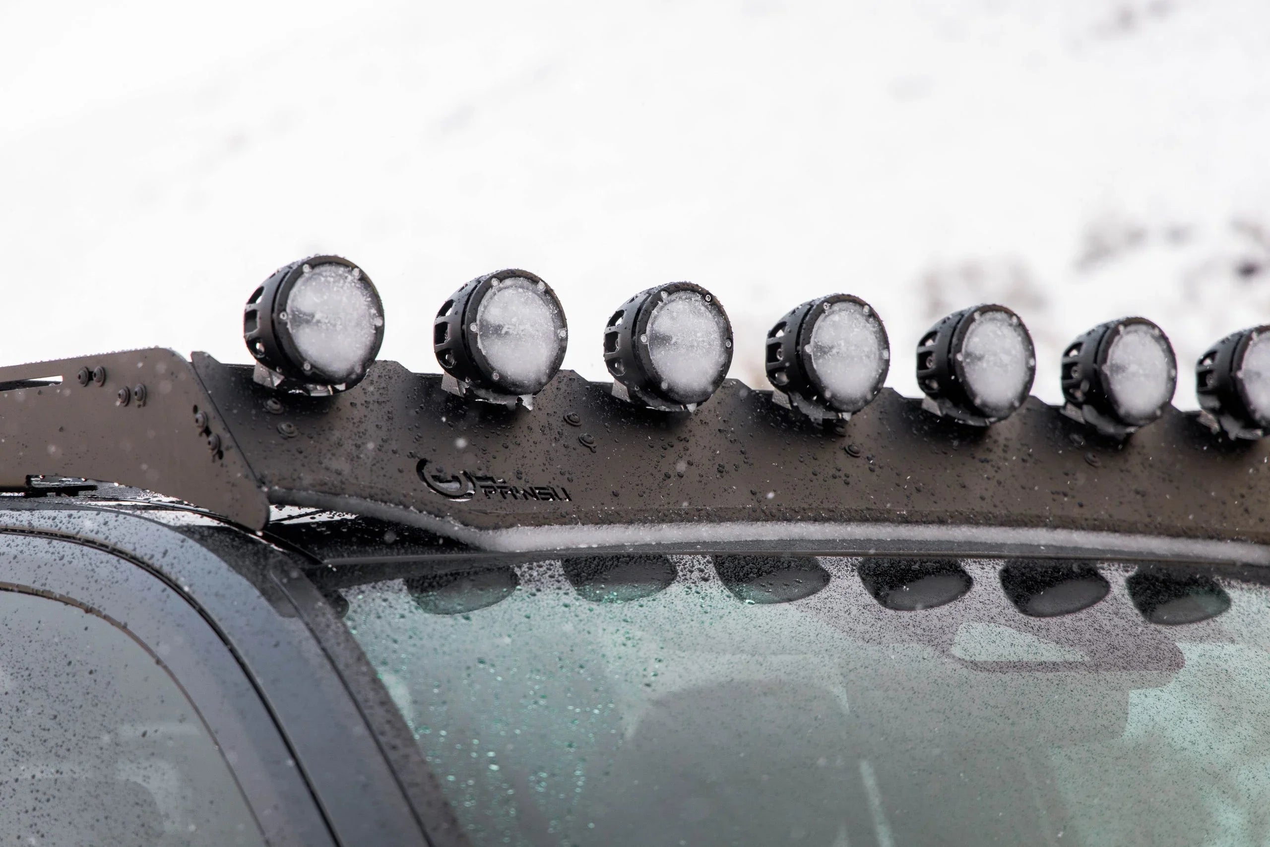 Close-up of Tacoma roof rack with round off-road LED lights, covered in snow, on a wet windshield.