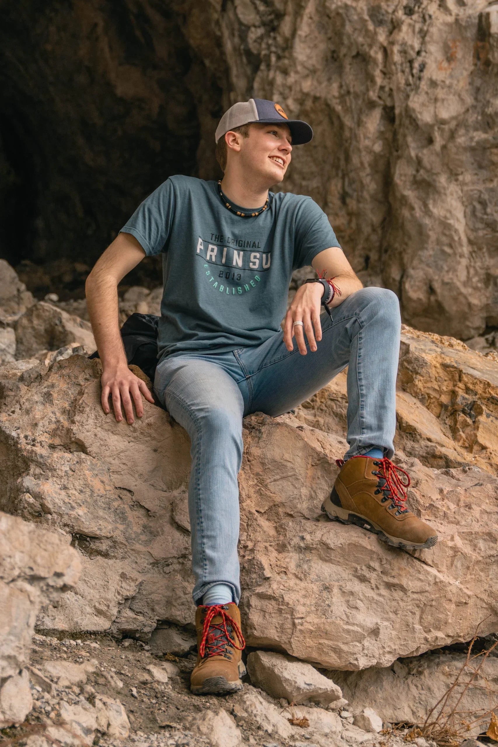 Man in blue Prinsu graphic shirt, jeans, and hiking boots sitting on rocky terrain outdoors