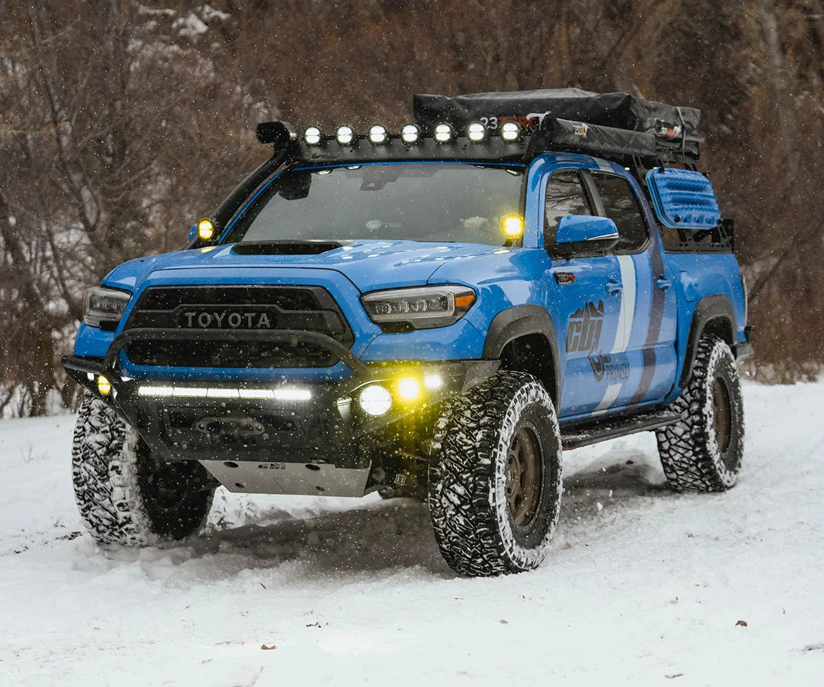 Blue Toyota Tacoma overland truck with rock sliders and camping gear driving in snow