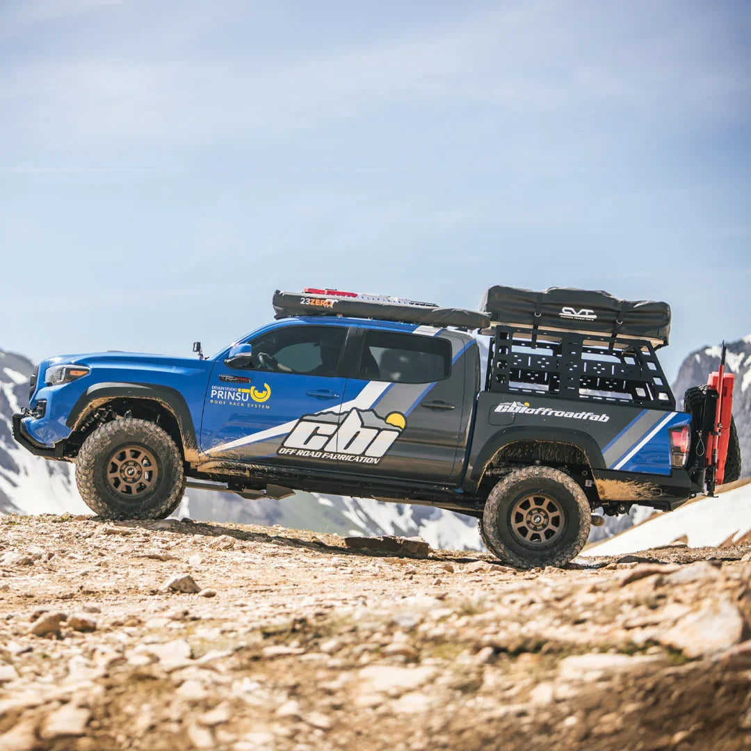 Blue Toyota Tacoma with roof rack and bed rack off-roading in a mountain landscape