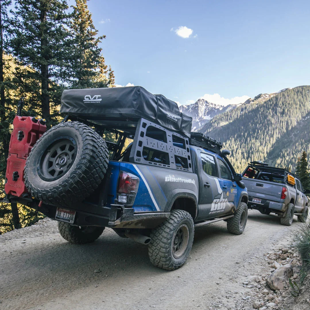 Toyota Tacoma trucks with roof and bed racks on rugged mountain trail, overlanding gear visible.