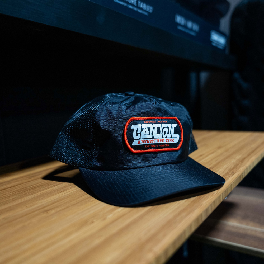 Black mesh trucker hat with 'Canyon Adventure Vans' patch on a wooden surface