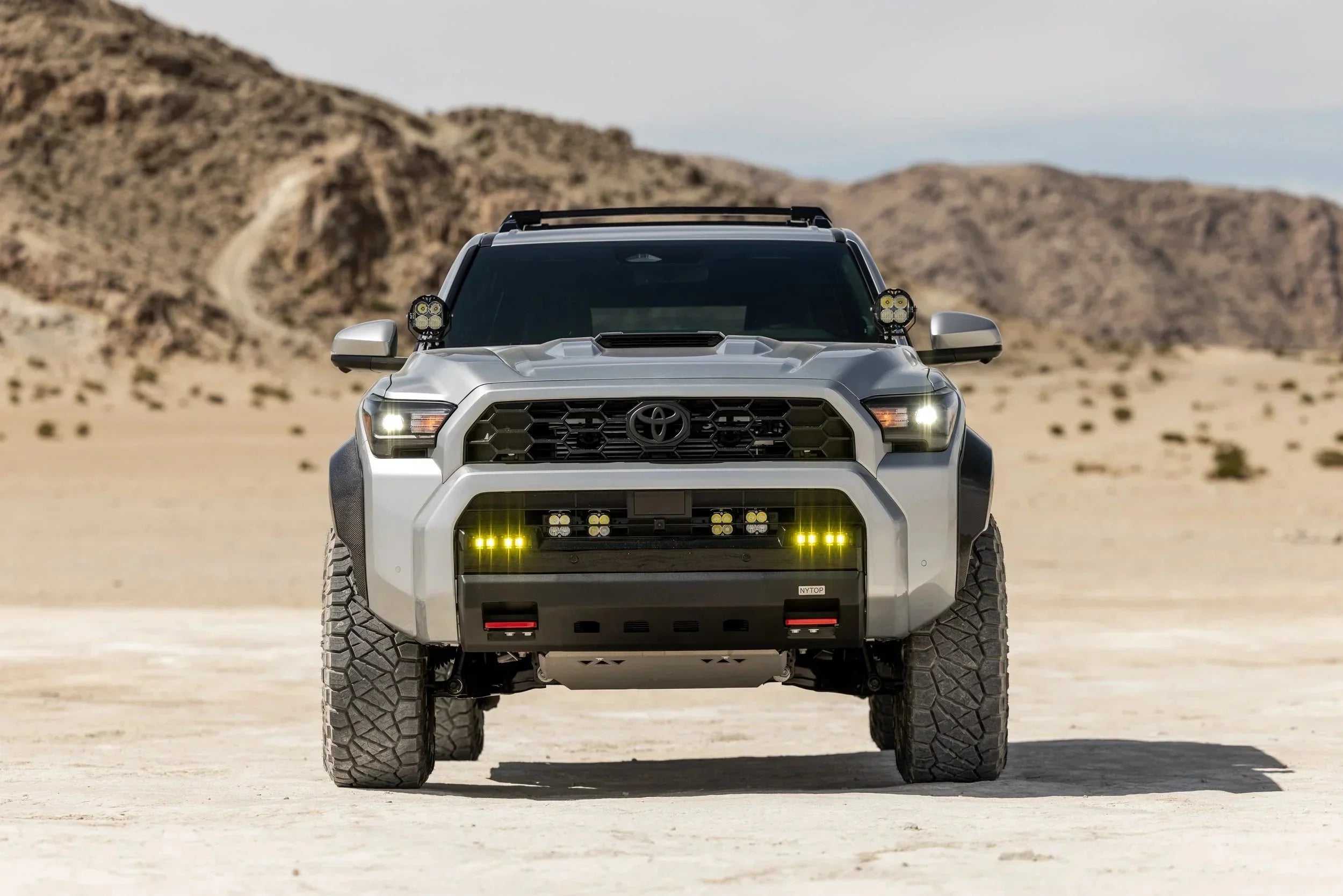 Front view of a silver Toyota Tacoma with off-road lights and rugged tires in a desert landscape