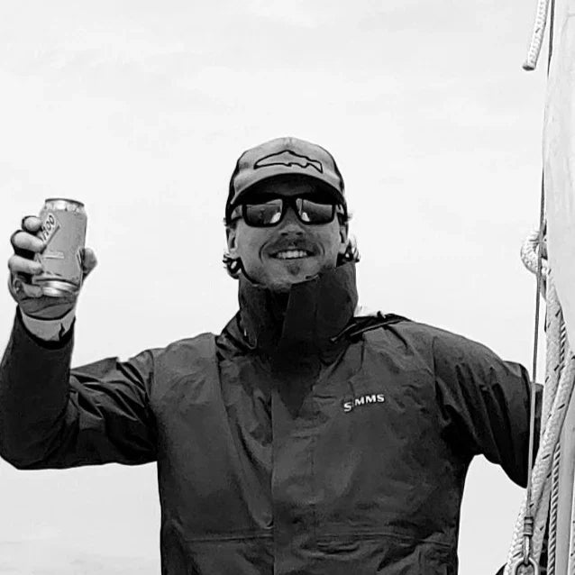 Man in outdoor gear and sunglasses holding a drink, wearing a cap, likely on an adventure trip
