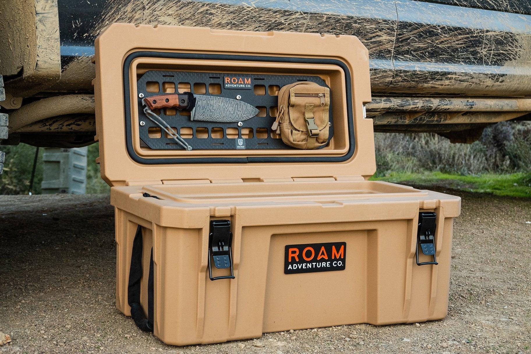 Tan overlanding storage box with knife and pouch, set outdoors beside a muddy off-road vehicle