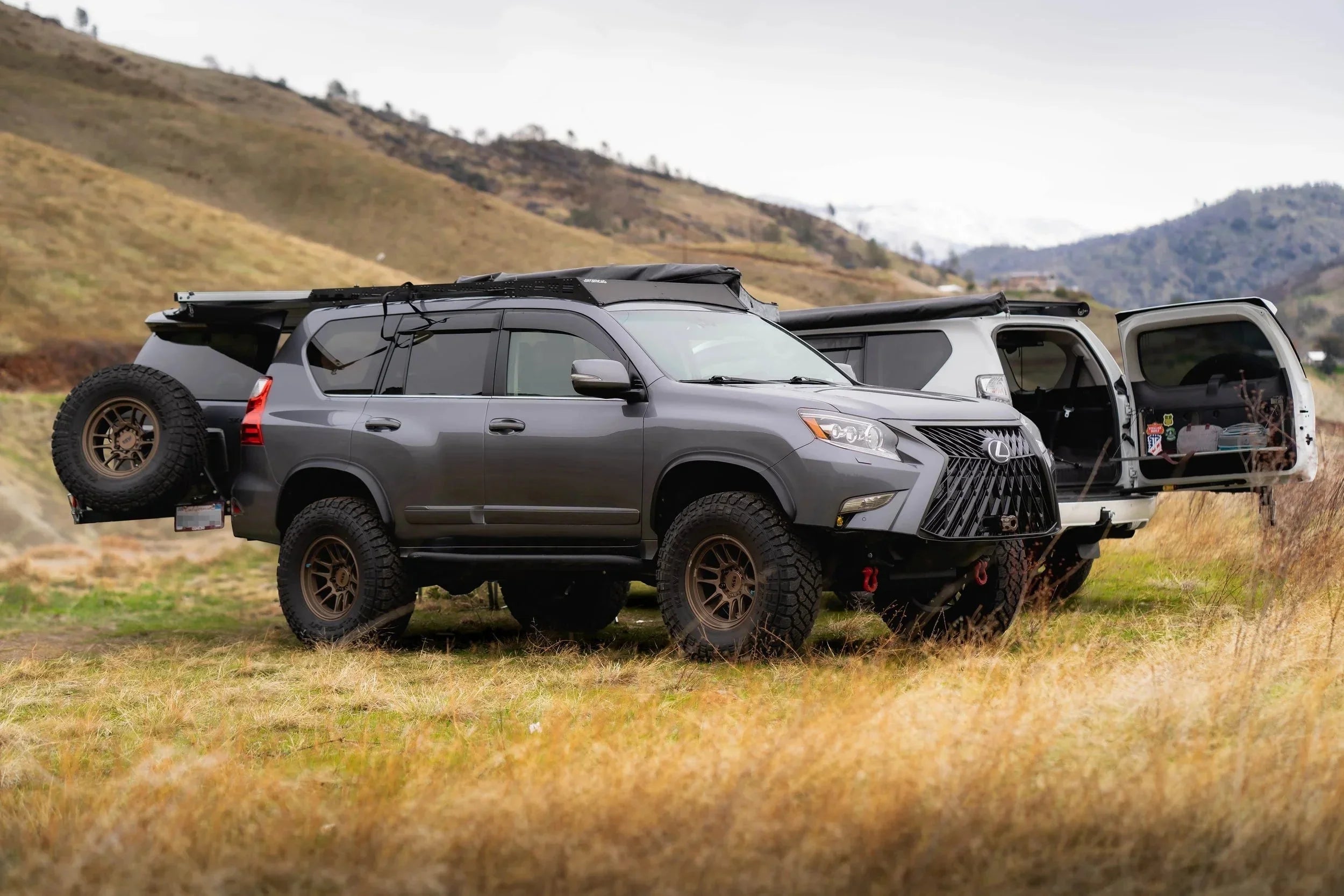 Lifted Lexus GX overland SUV with roof rack and spare tire, parked in grassy hills, expedition camping setup