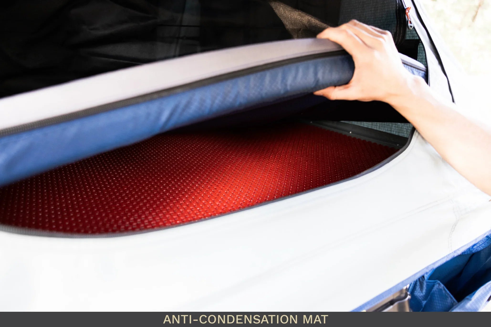 Hand lifting rooftop tent mattress to reveal red anti-condensation mat inside camping setup
