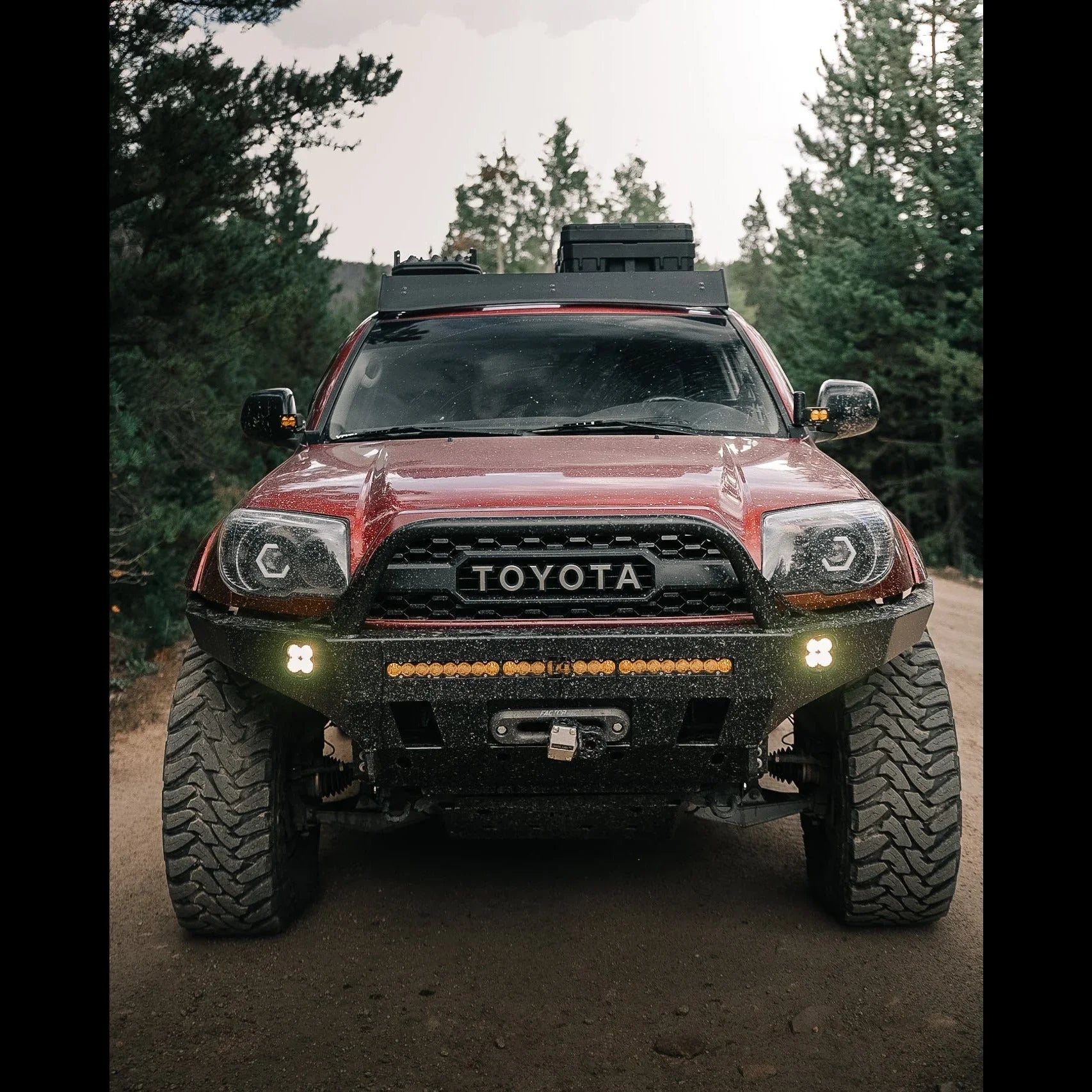 Red Toyota Tacoma with off-road bumper, roof rack, and LED lights on a forest trail