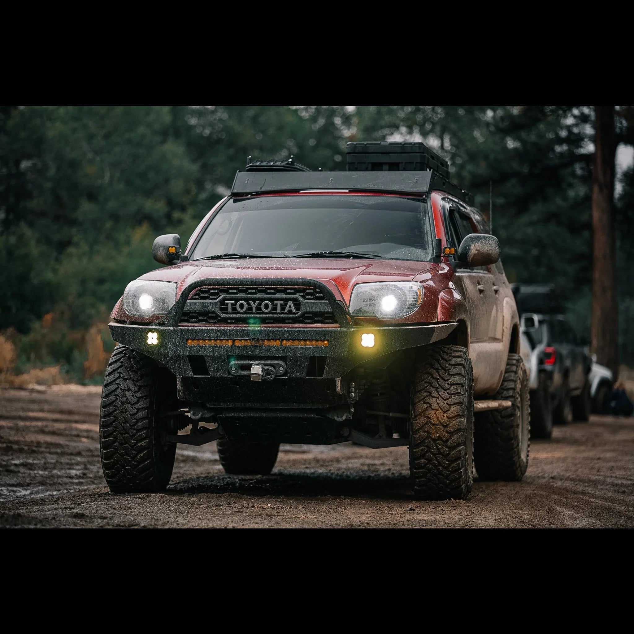 Red Toyota Tacoma overland vehicle with off-road tires, rooftop gear, forest trail