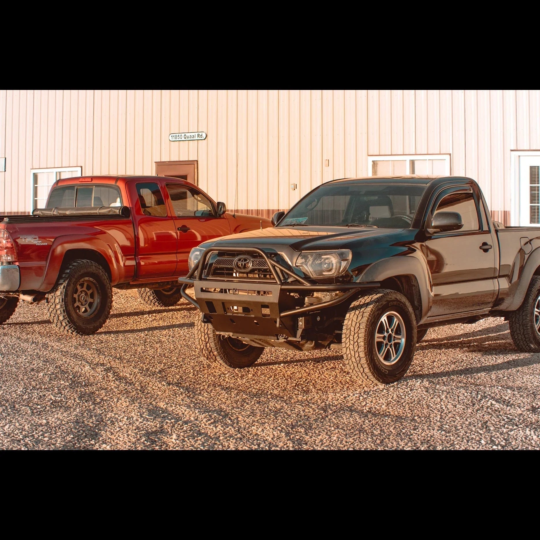 Lifted Toyota Tacoma trucks with off-road bumpers parked on gravel outside a metal building