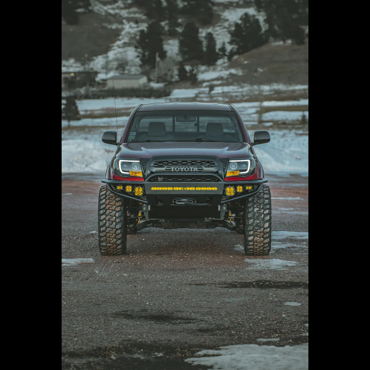 Front view of modified Toyota Tacoma with off-road bumper and LED lights in snowy terrain