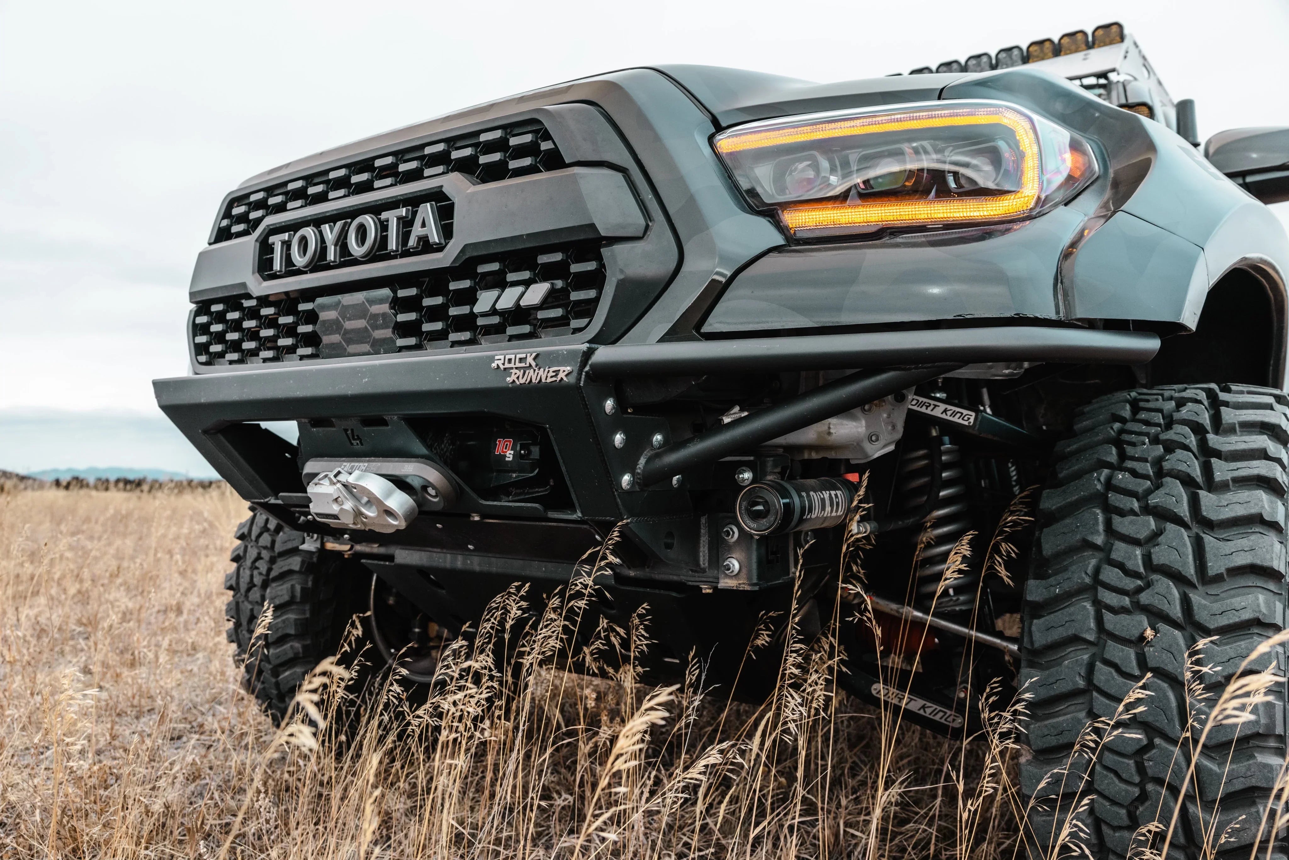 Toyota Tacoma off-road front bumper with winch, LED lights, and oversized all-terrain tires in grass field