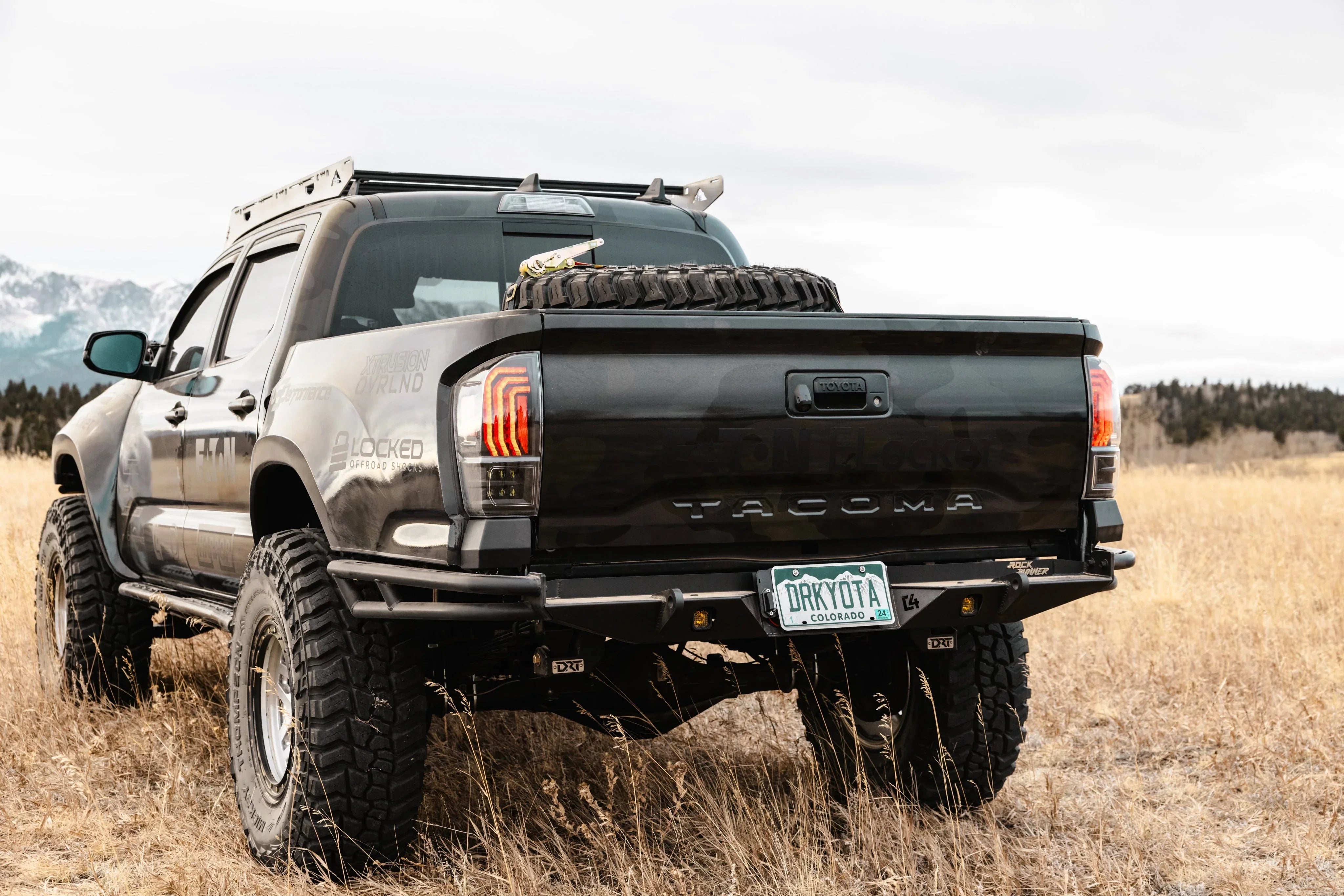 Lifted Toyota Tacoma truck with off-road tires, spare tire, and roof rack in a grassy field.