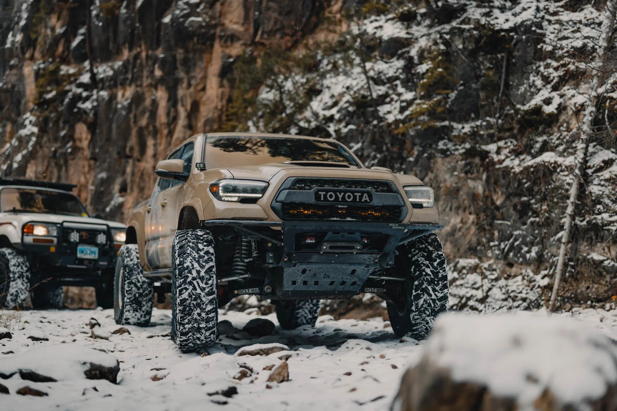 Lifted Toyota Tacoma with off-road tires and accessories on a snowy, rocky trail in the mountains
