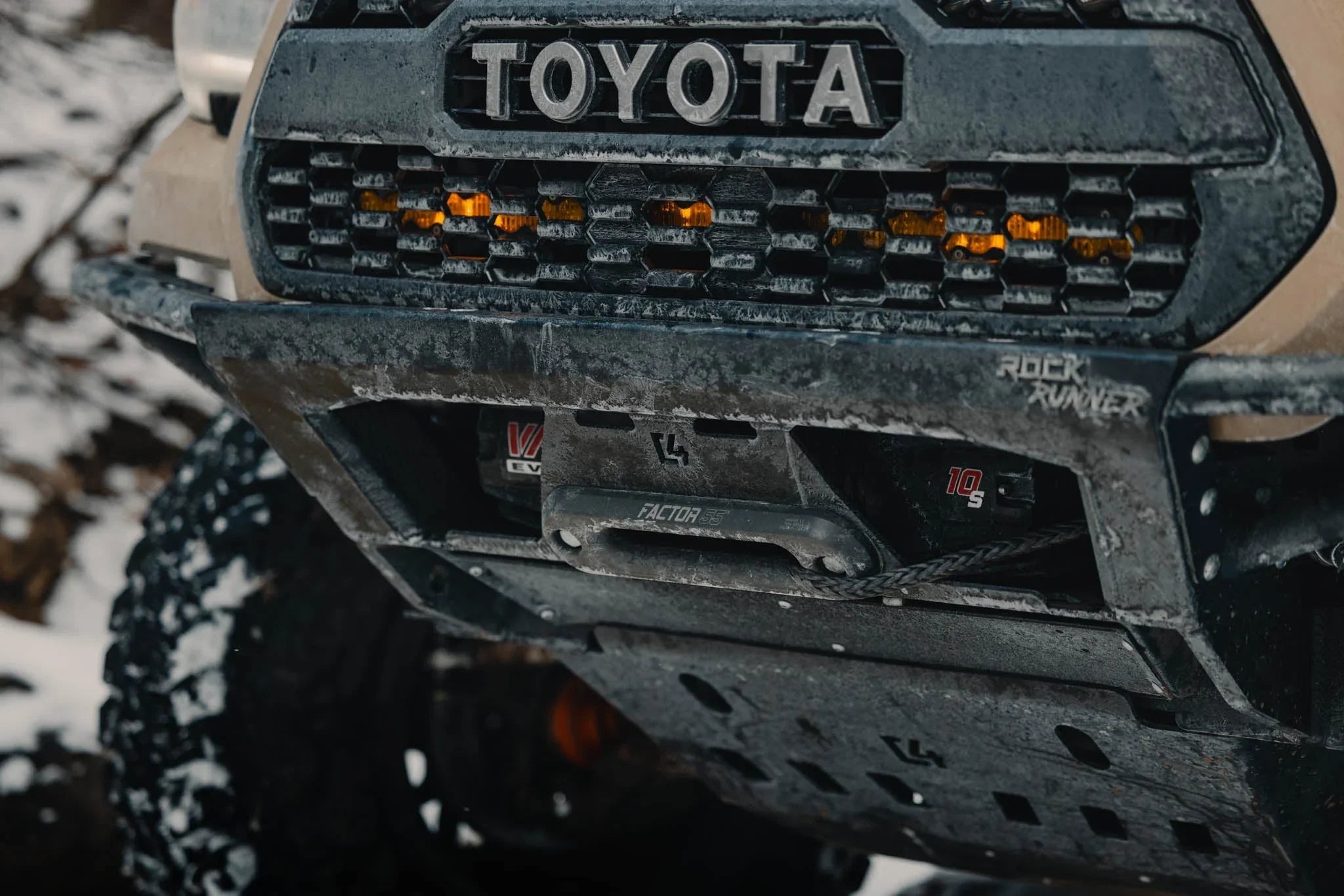 Close-up of Toyota Tacoma off-road front bumper with winch and skid plate in snowy terrain.