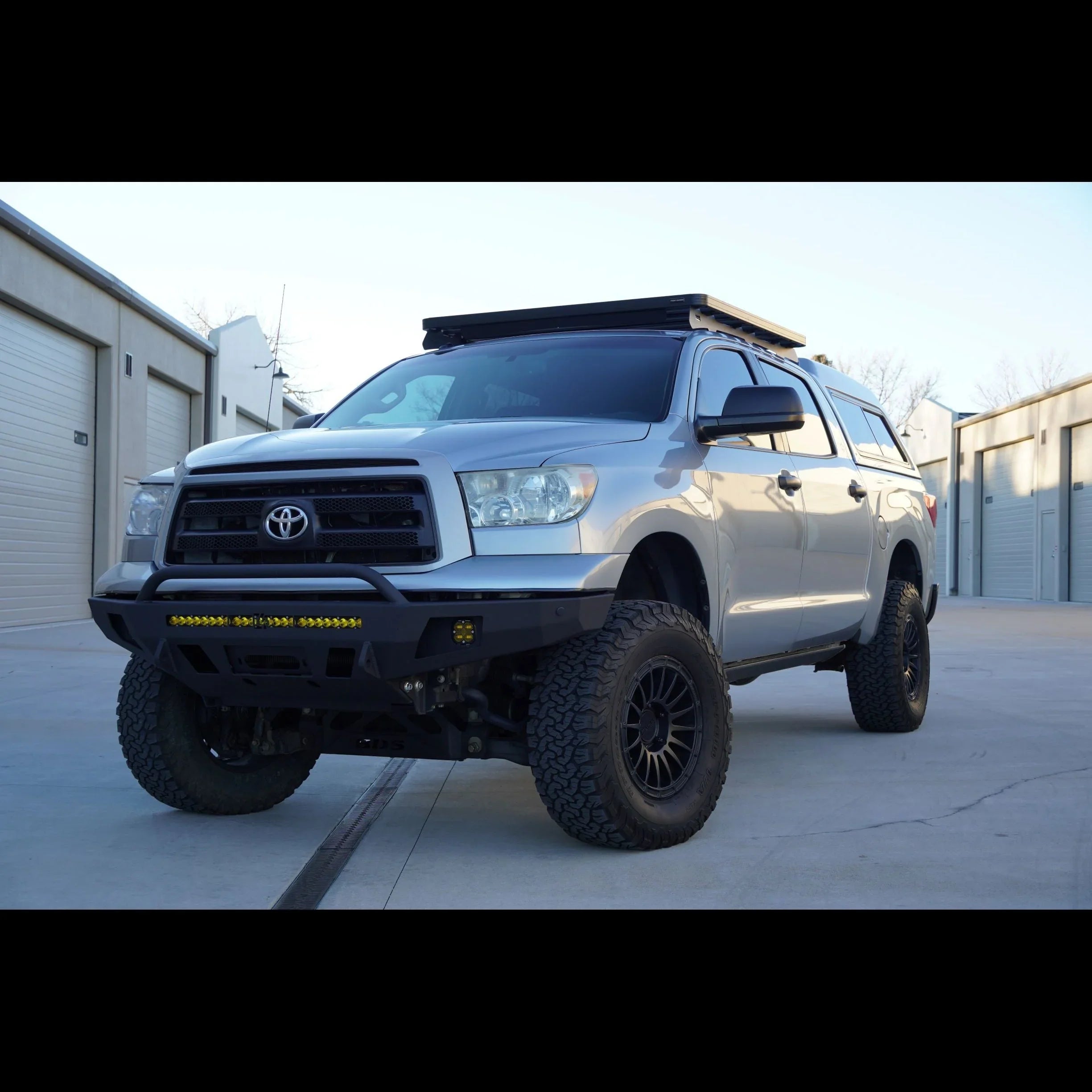 Lifted silver Toyota Tundra with off-road bumper, roof rack, and all-terrain tires in driveway