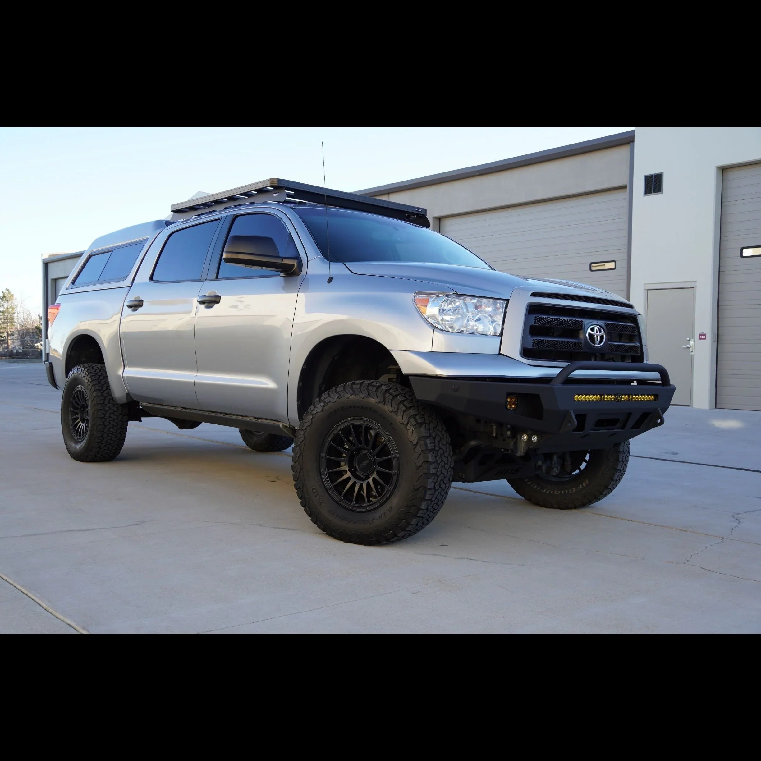 Lifted silver Toyota Tundra with off-road tires, aftermarket bumper, roof rack, and camper shell
