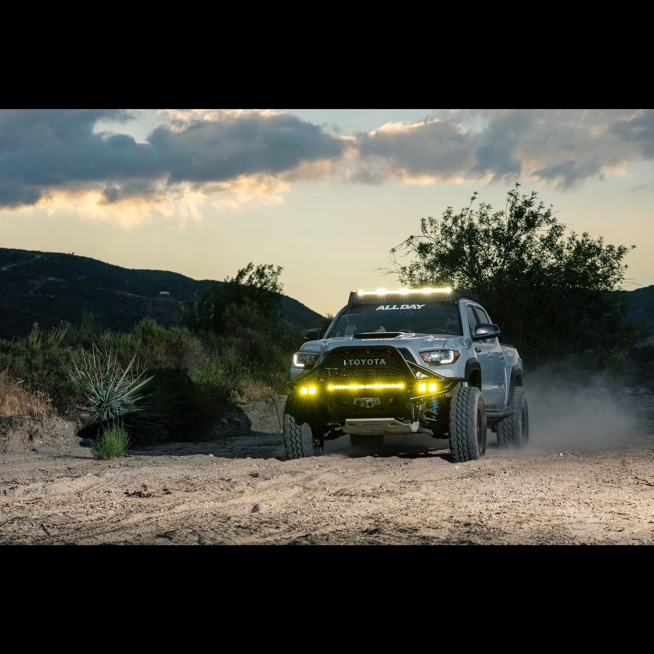 Toyota Tacoma equipped with off-road LED lights driving on a rugged dirt trail at dusk
