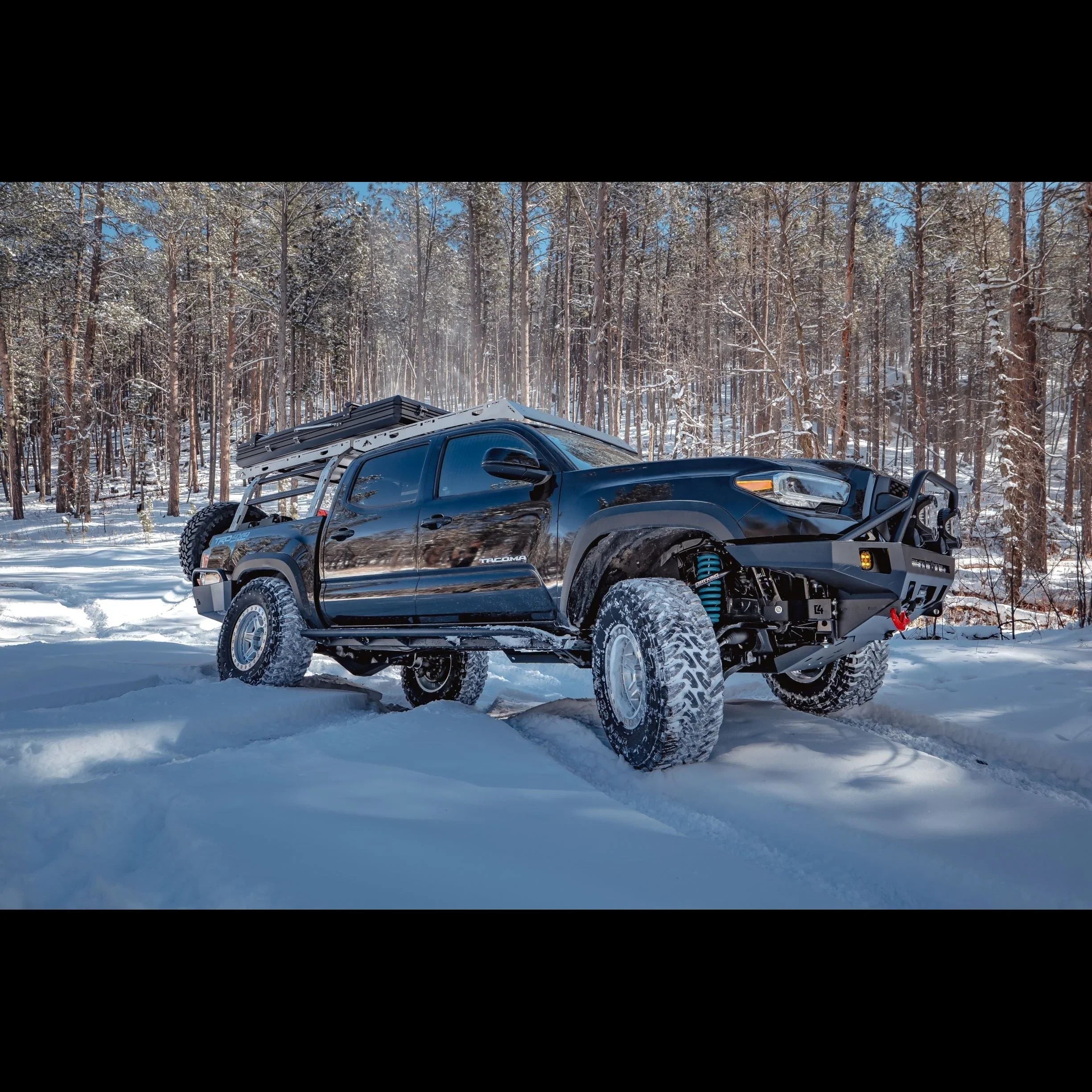 Toyota Tacoma with overland front bumper driving in snowy forest, off-road gear visible