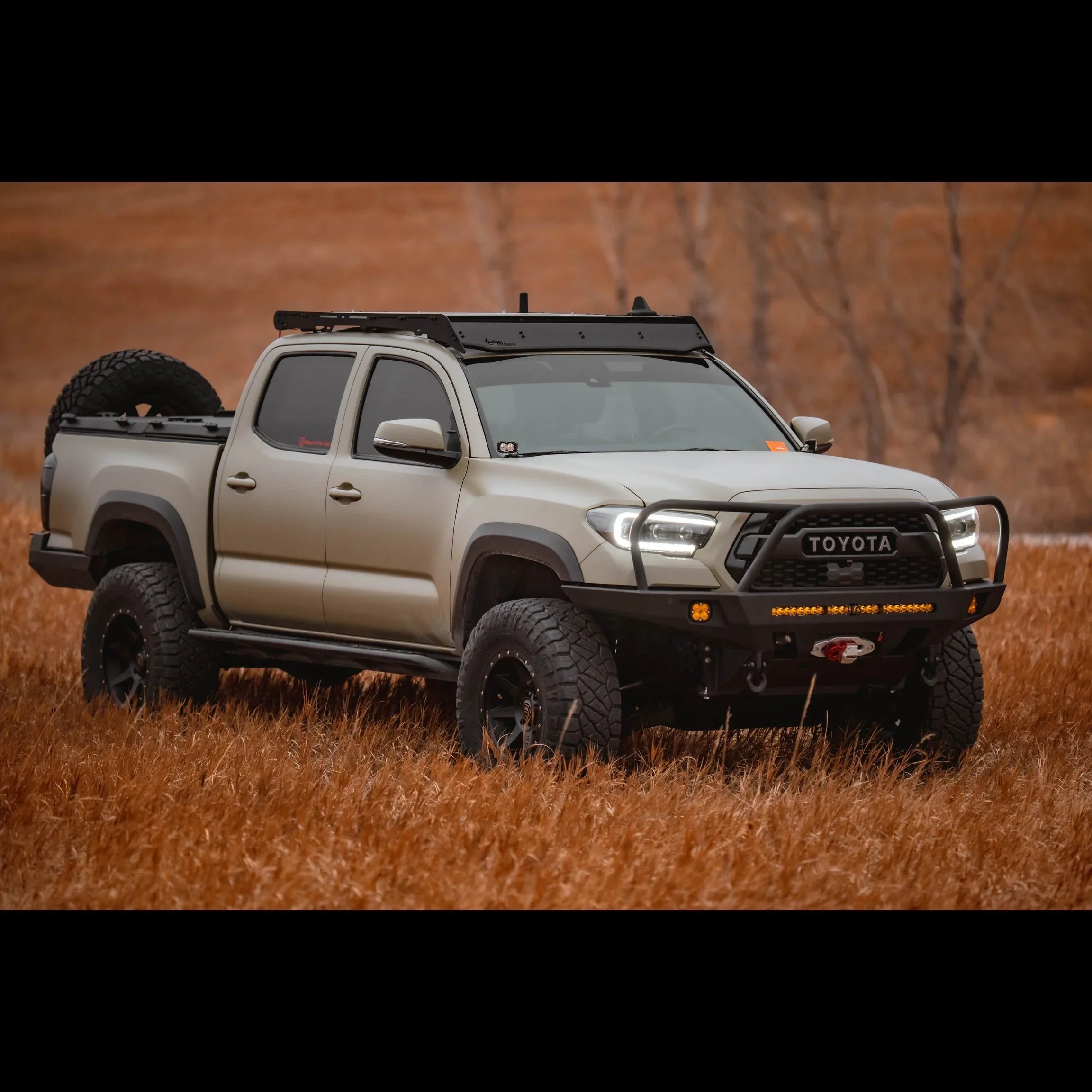 Lifted 3rd gen Toyota Tacoma with overland front bumper, roof rack, and off-road tires in a field