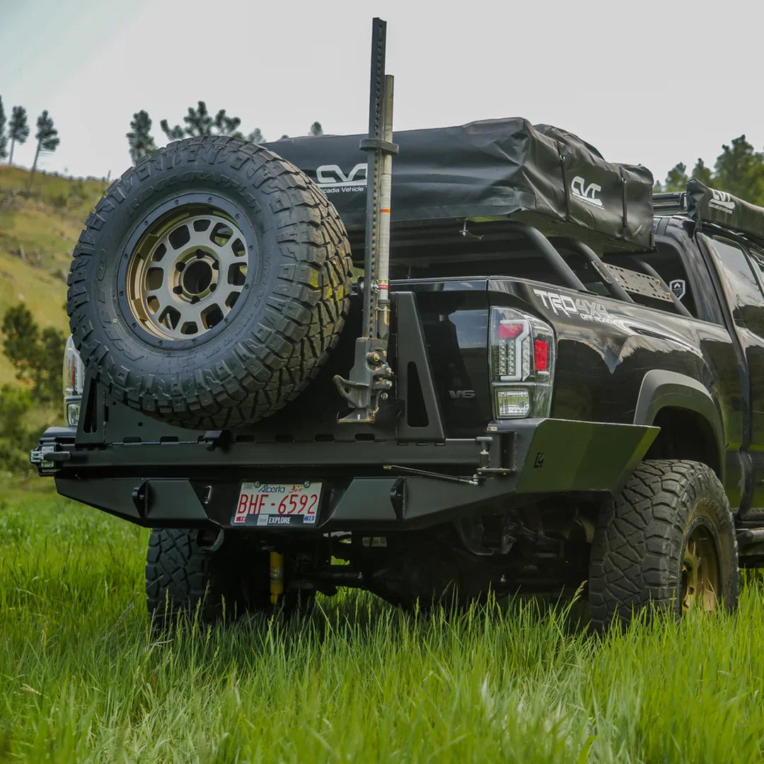 Toyota Tacoma with overland high clearance rear bumper, spare tire, and camping gear in grassy field