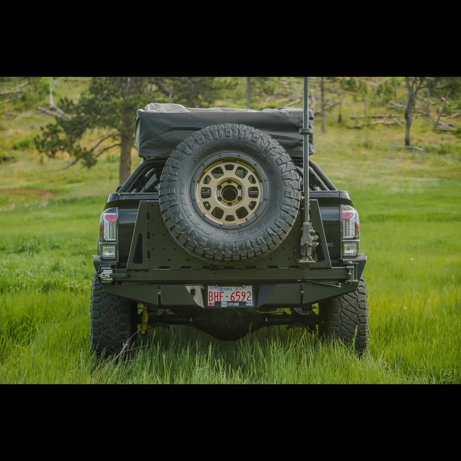 Toyota Tacoma with overland rear bumper, spare tire mount, and camping gear in grassy field