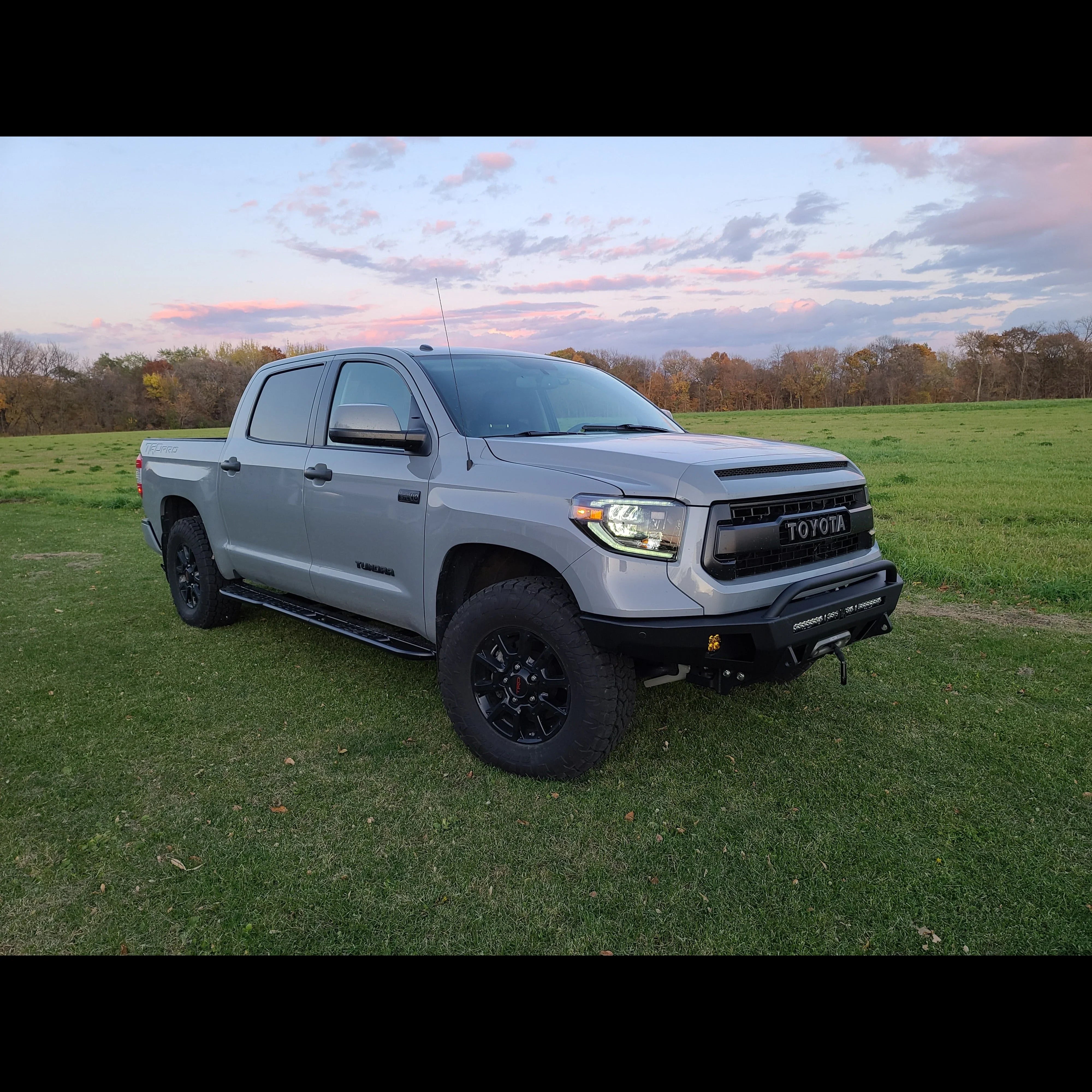 Gray Toyota Tundra with off-road tires and custom bumper parked on grassy field at sunset