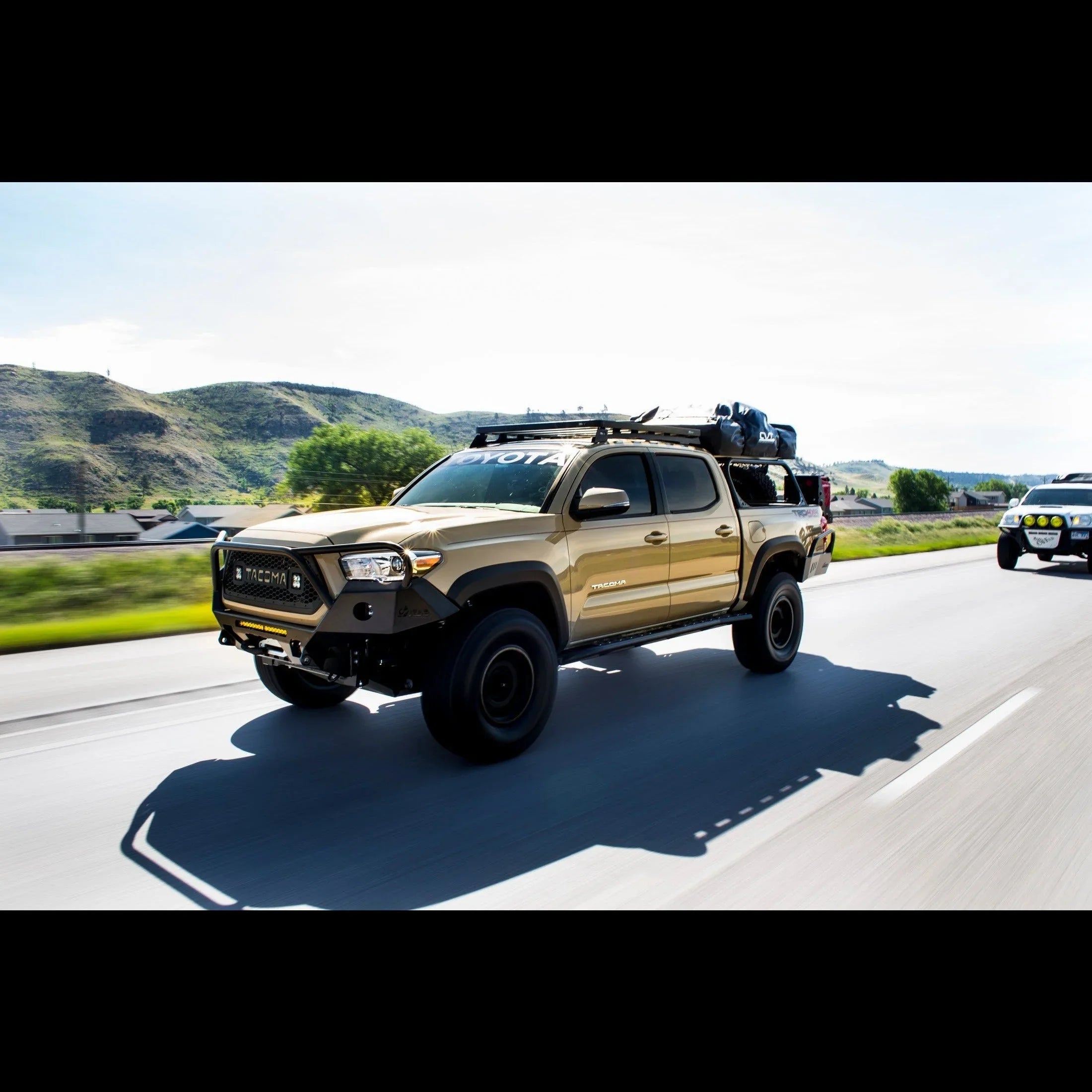 Tan Toyota Tacoma overland truck with camping gear driving on highway in scenic mountain area
