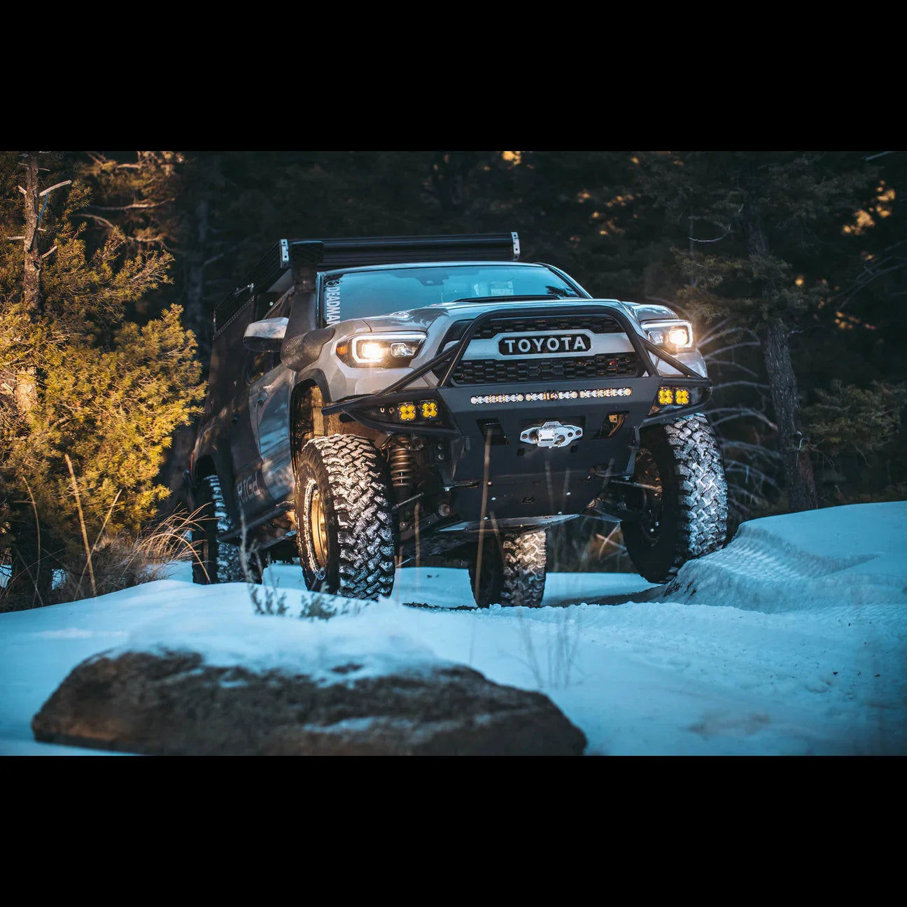 Lifted Toyota Tacoma with off-road bumper and lights driving on snowy trail in pine forest