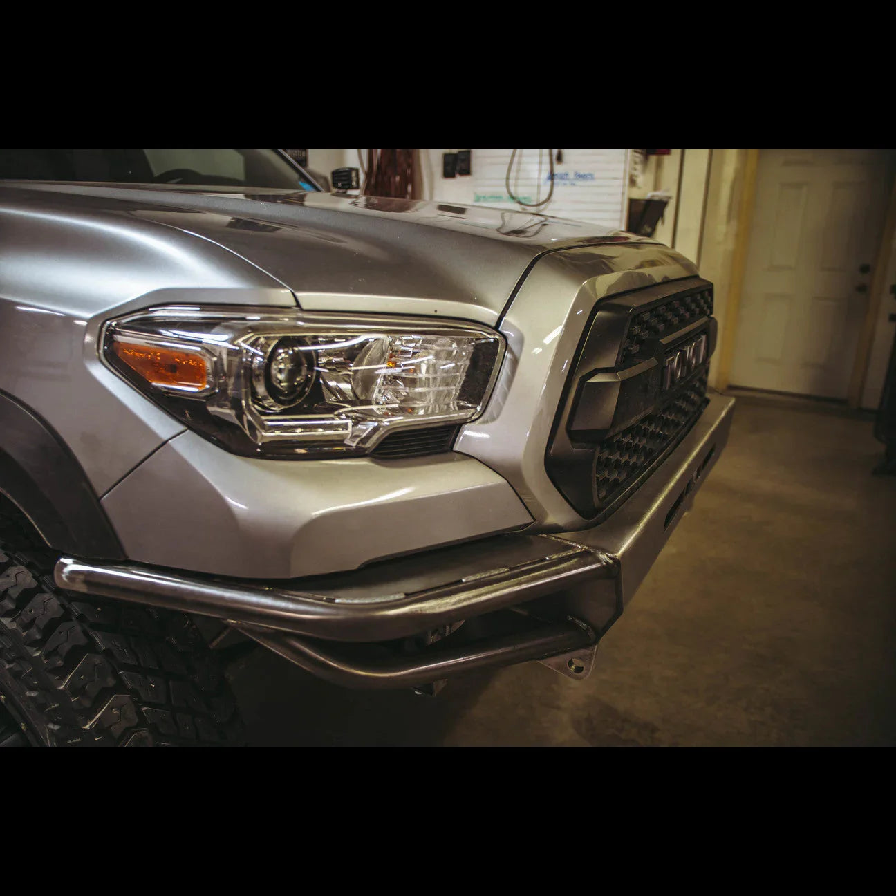 Front view of a silver Toyota Tacoma with custom off-road steel bumper in a garage.