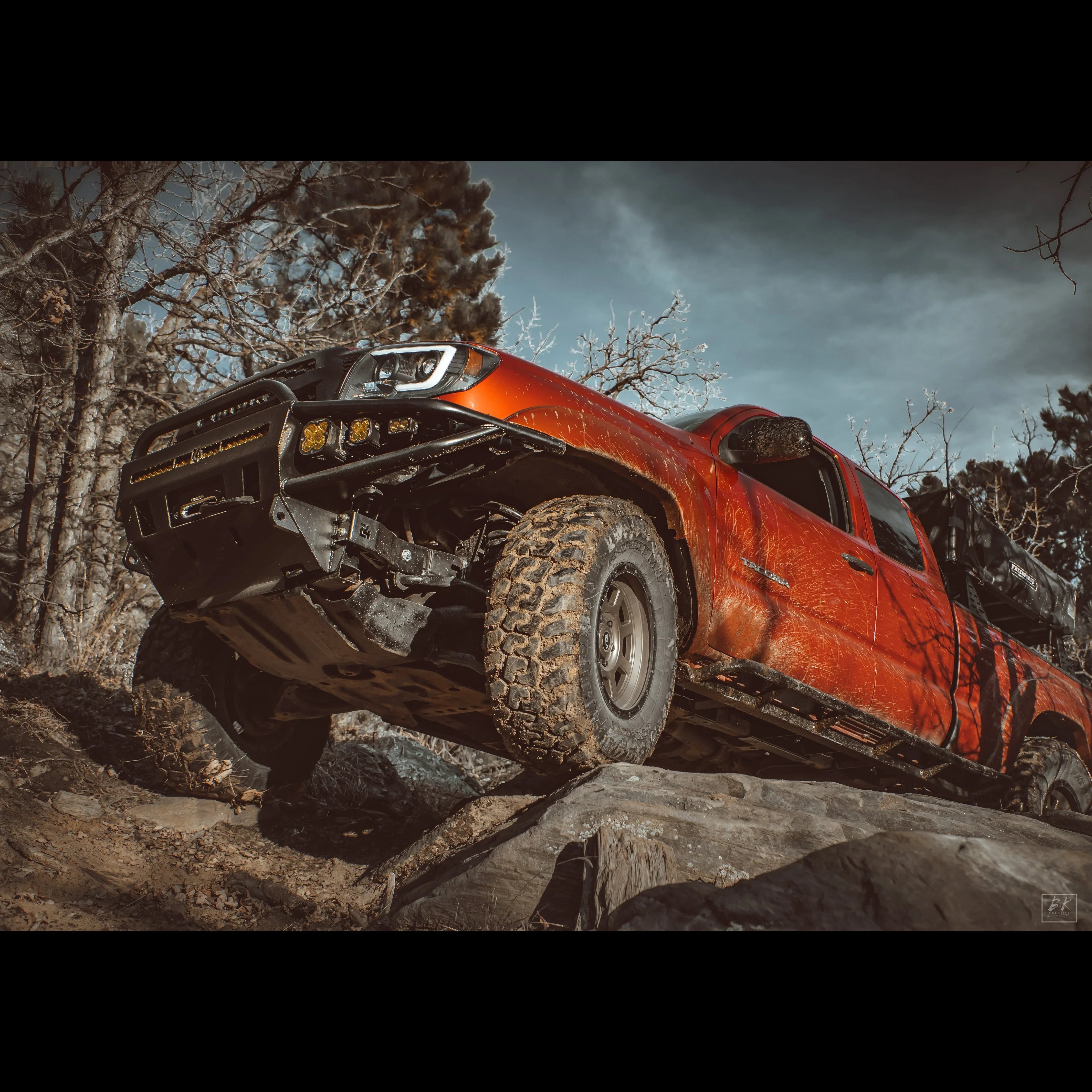 Toyota Tacoma overland truck with off-road accessories driving on rocky terrain in the woods
