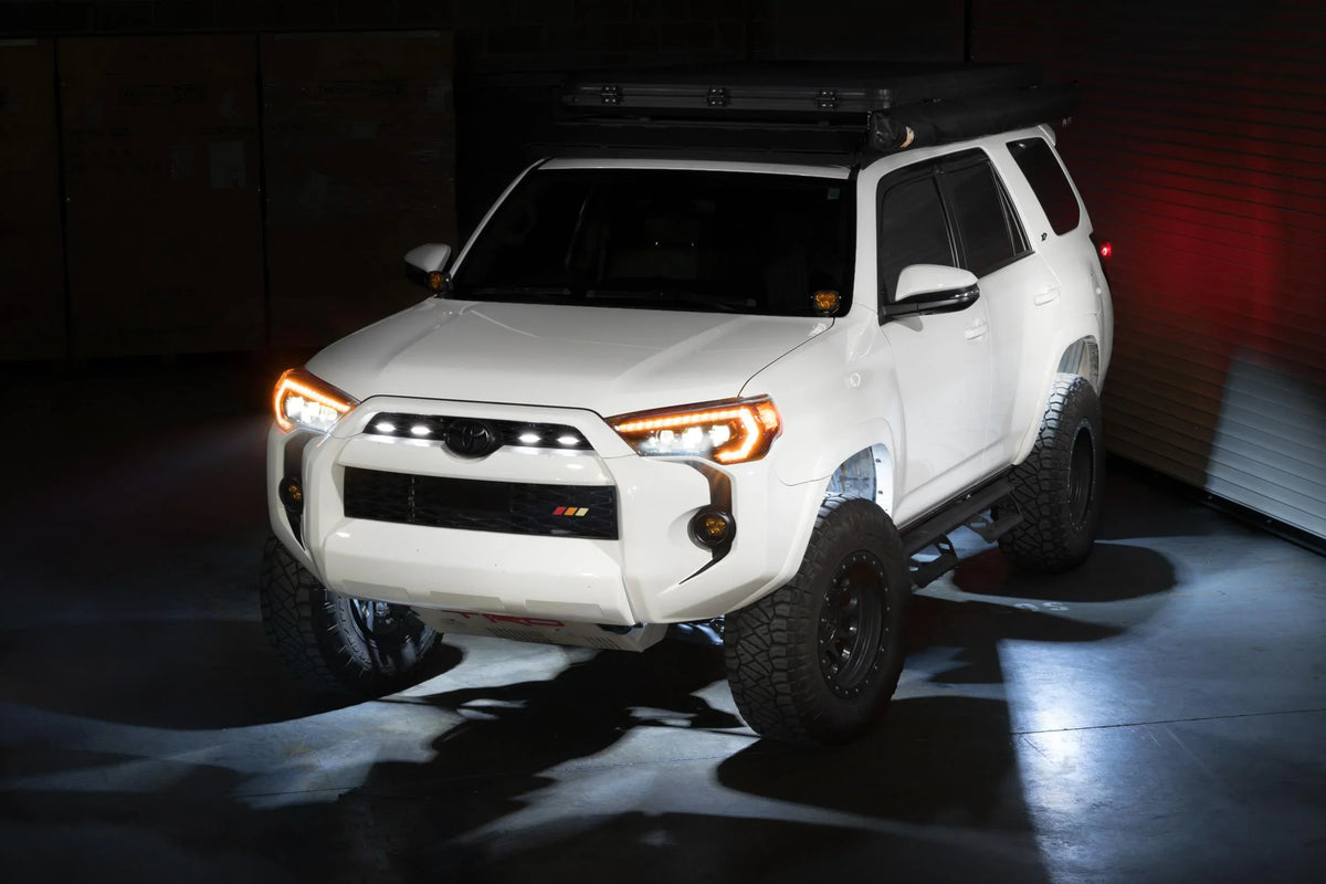 White Toyota 4Runner with off-road tires, roof rack, and LED lights in a dim garage.