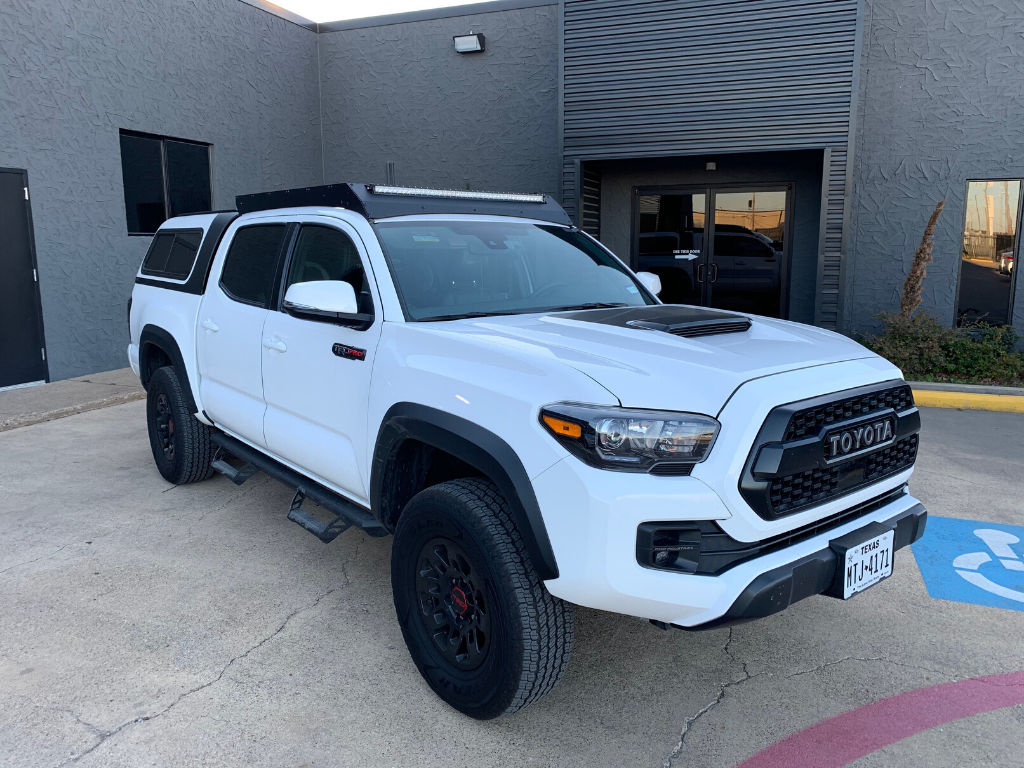 White Toyota Tacoma with roof rack and camper shell in urban setting, overlanding ready