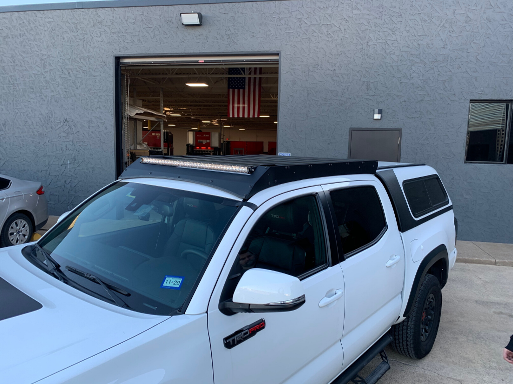 White Toyota Tacoma with roof rack and LED light bar, parked outside garage