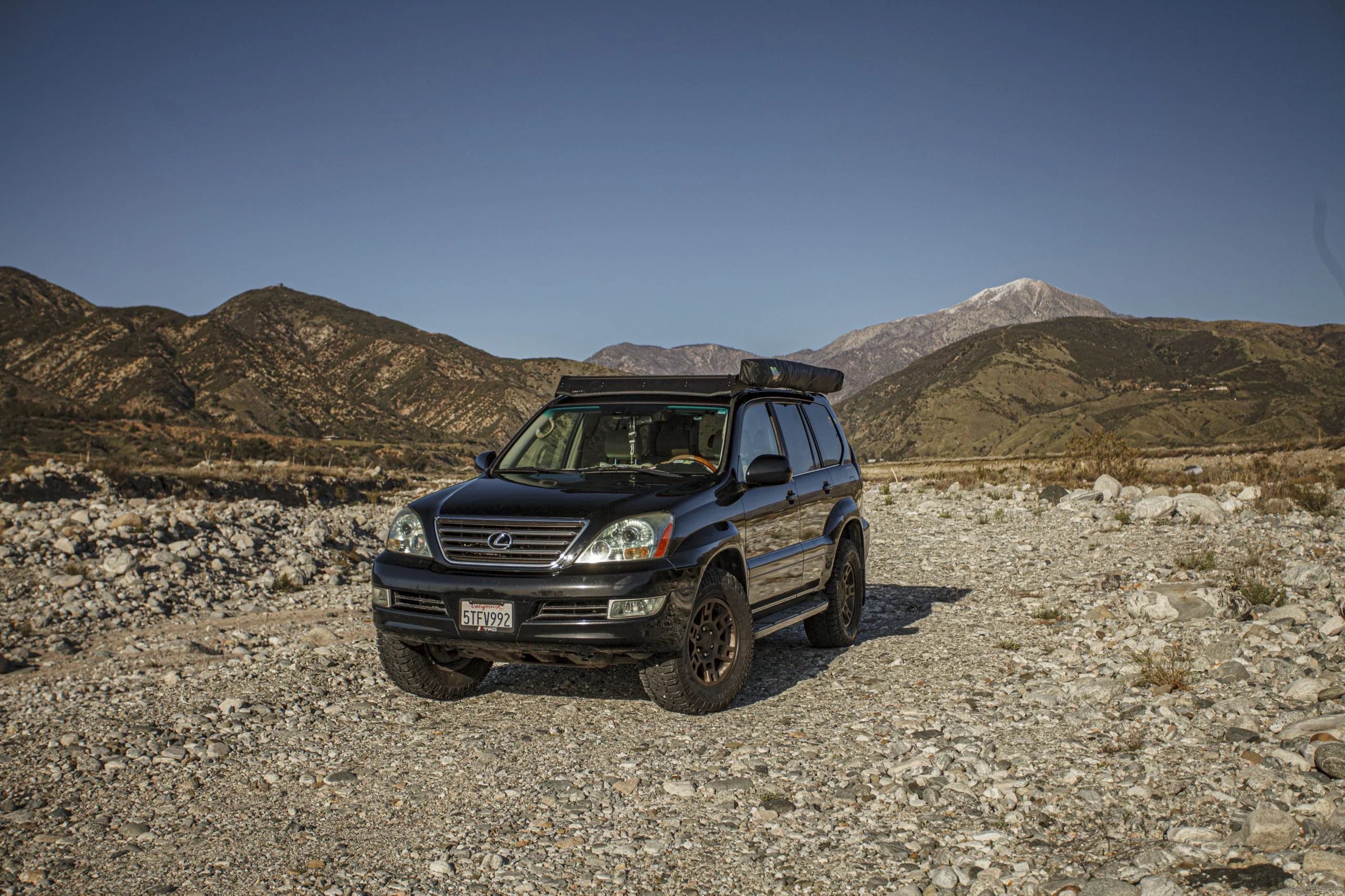 Black Lexus GX470 overland SUV with roof rack parked on rocky terrain, mountains in background