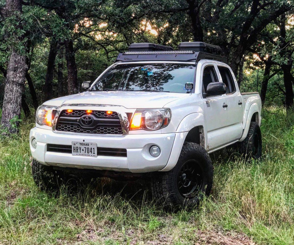 White second-gen Toyota Tacoma with Prinsu roof rack parked in a forest, off-road gear visible.