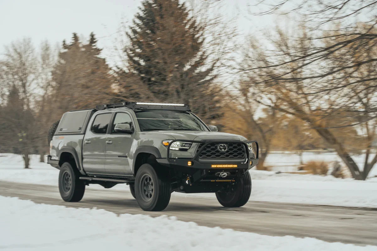 Toyota Tacoma with overlanding gear driving on a snowy road, equipped with LED lights and roof rack
