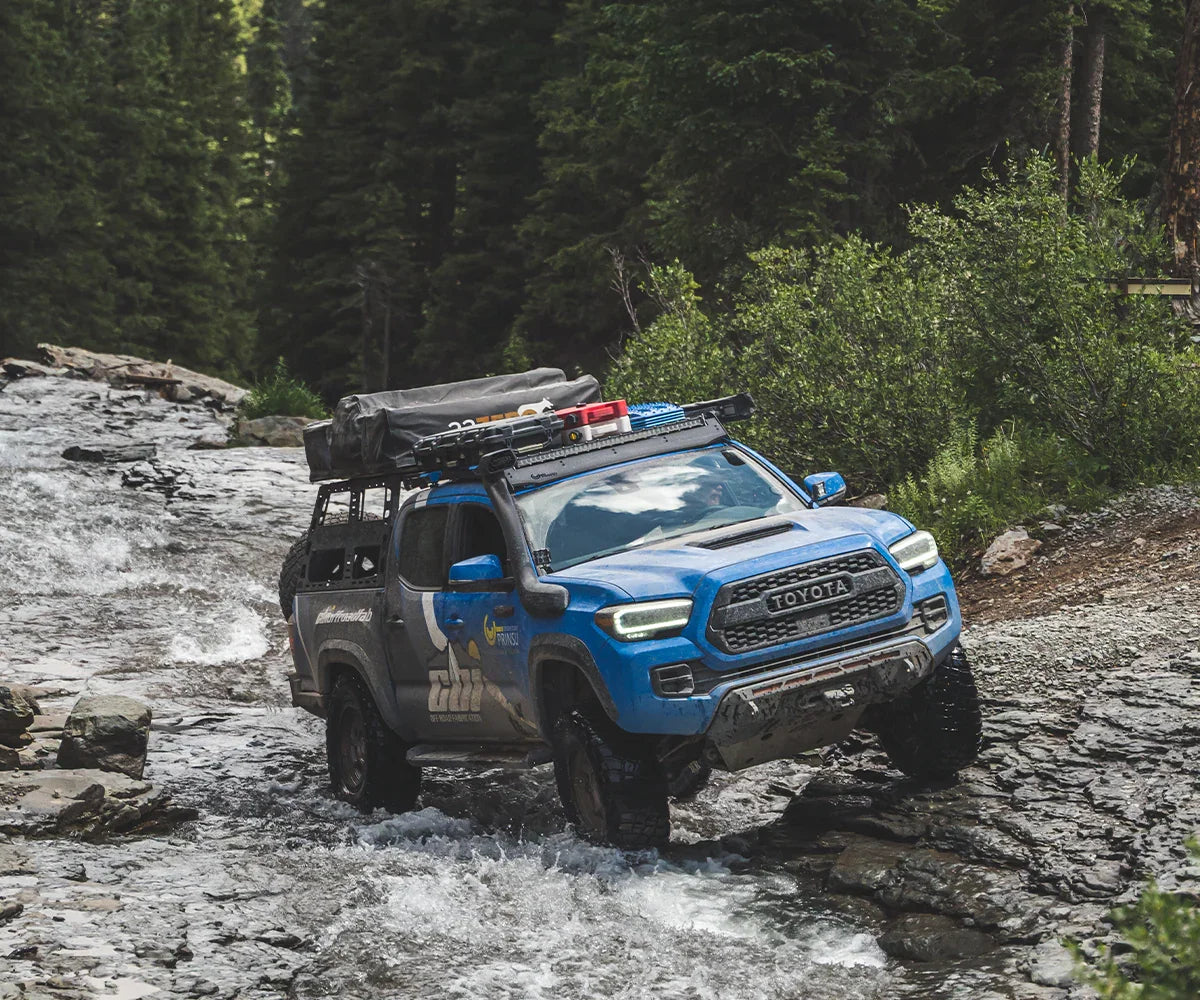 Blue Toyota Tacoma with front bumper and gear driving off-road through rocky creek