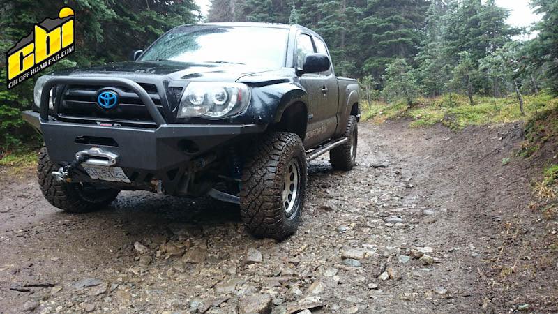 Toyota Tacoma with aftermarket aluminum off-road bumper and winch on a muddy forest trail.