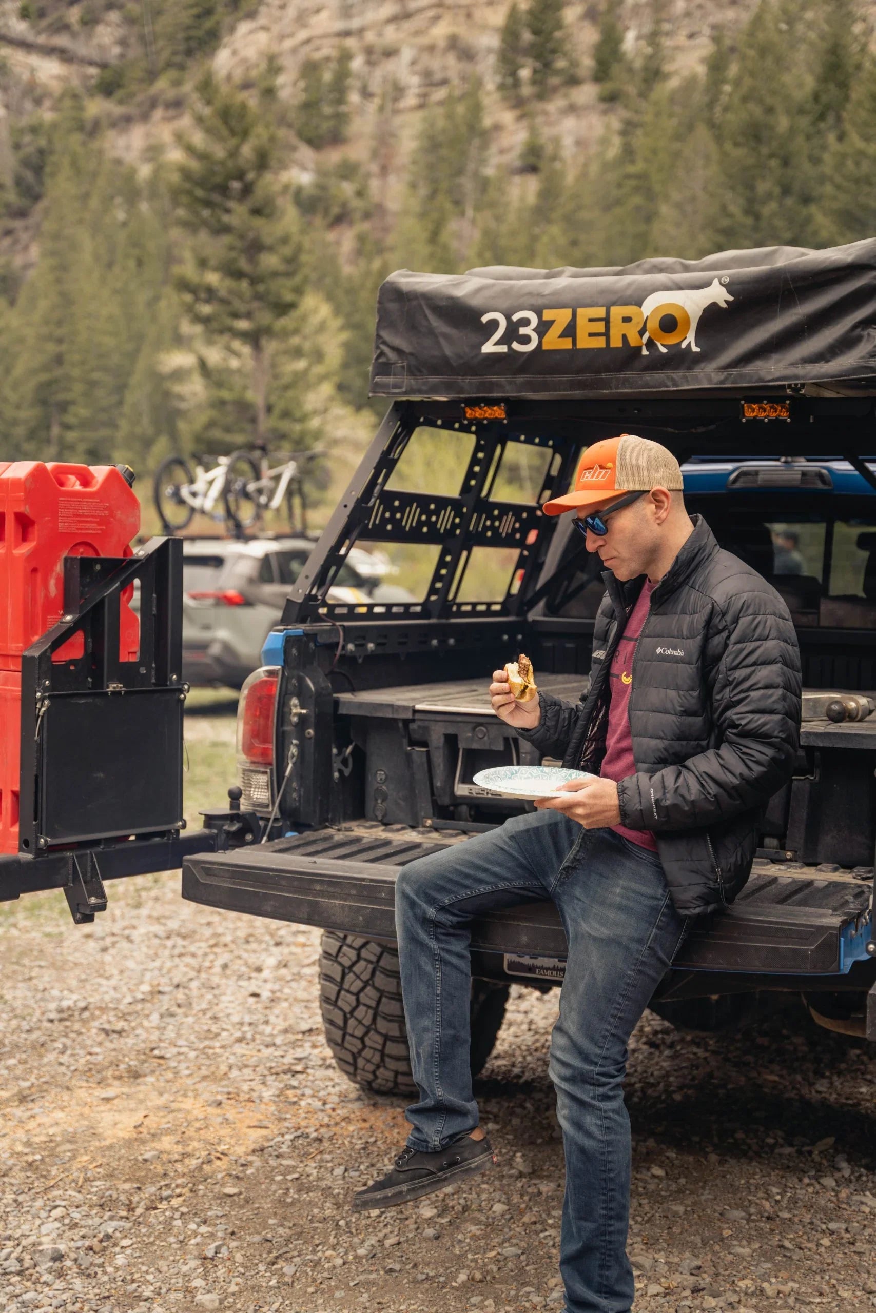 Man eating at camp table on off-road truck tailgate with camping gear in forest setting