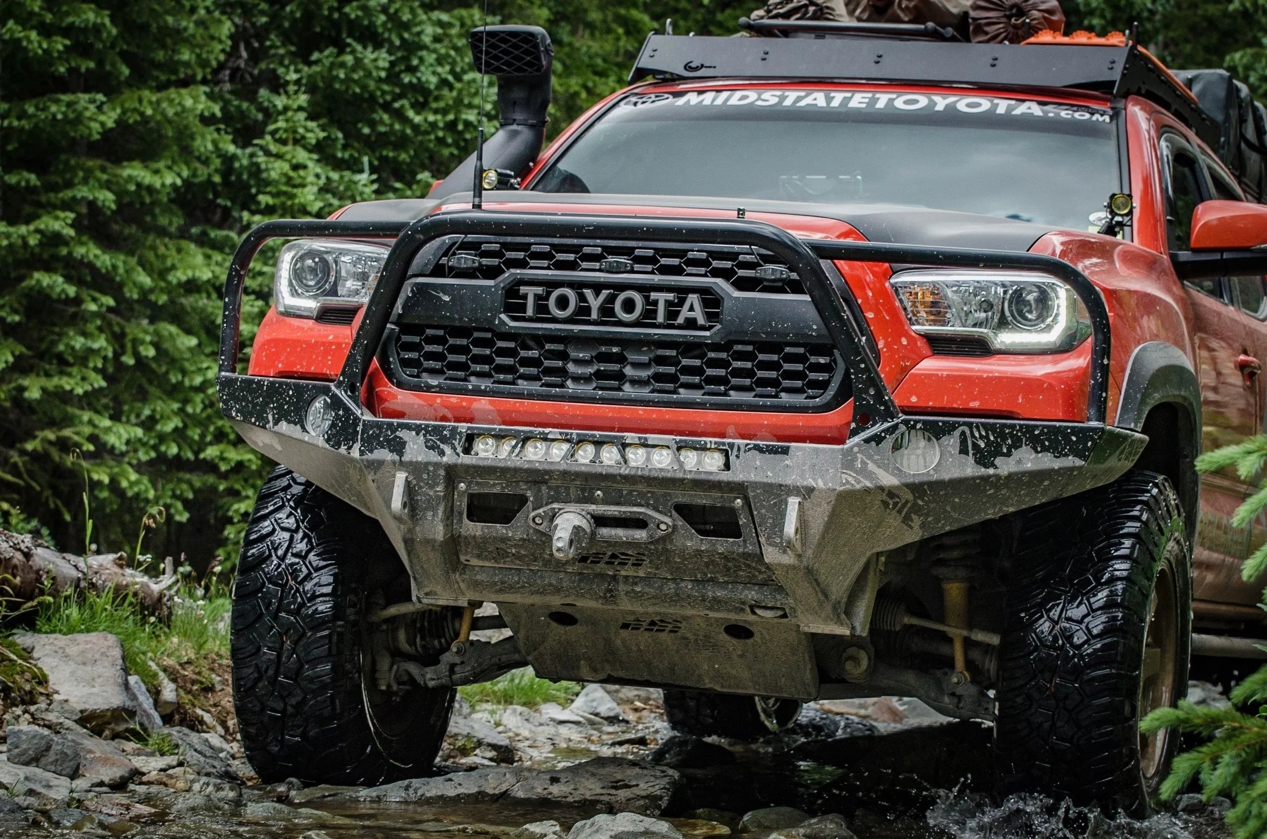 Toyota Tacoma with off-road front bumper driving through a rocky creek in a forest