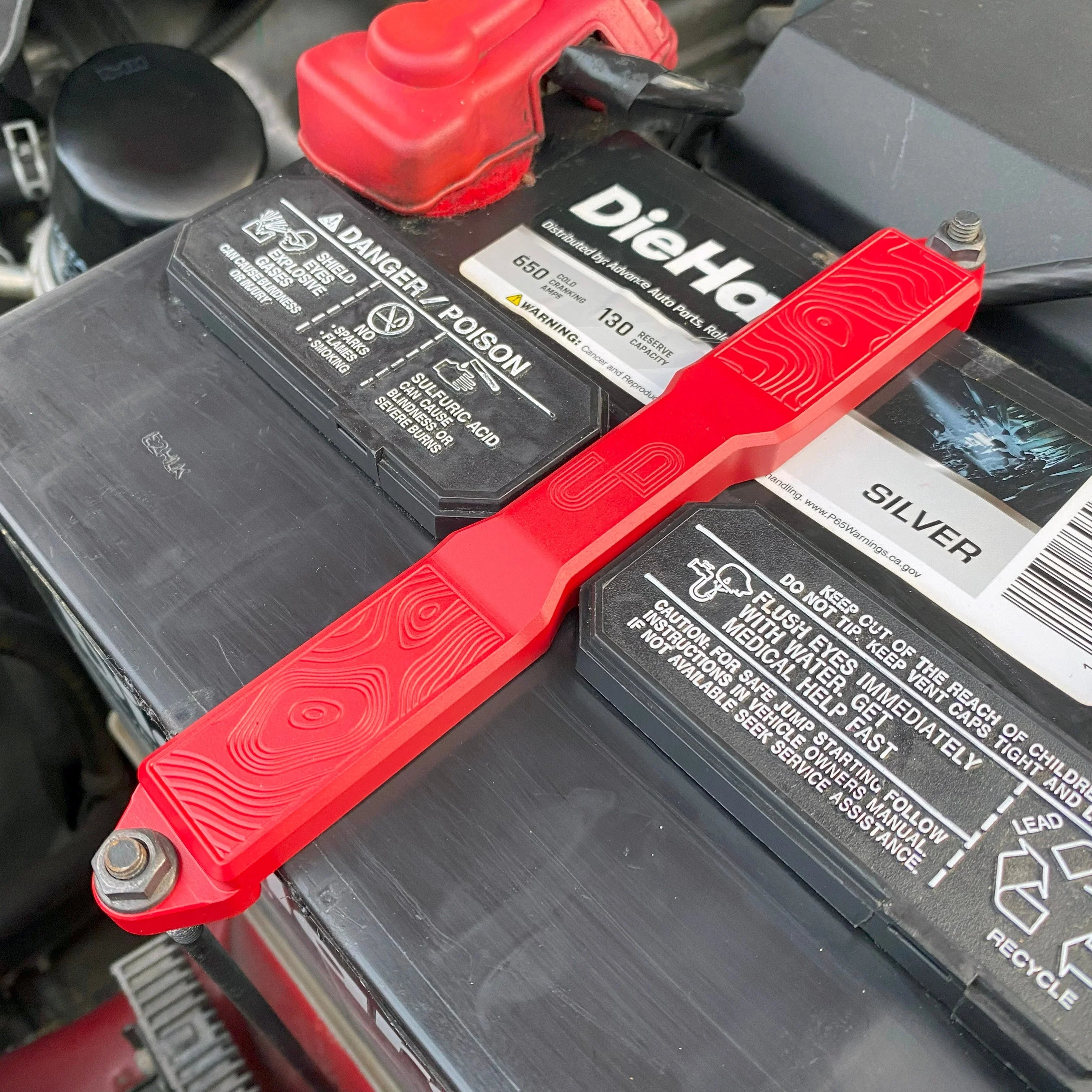 Red billet aluminum battery tie down installed on a DieHard car battery under vehicle hood