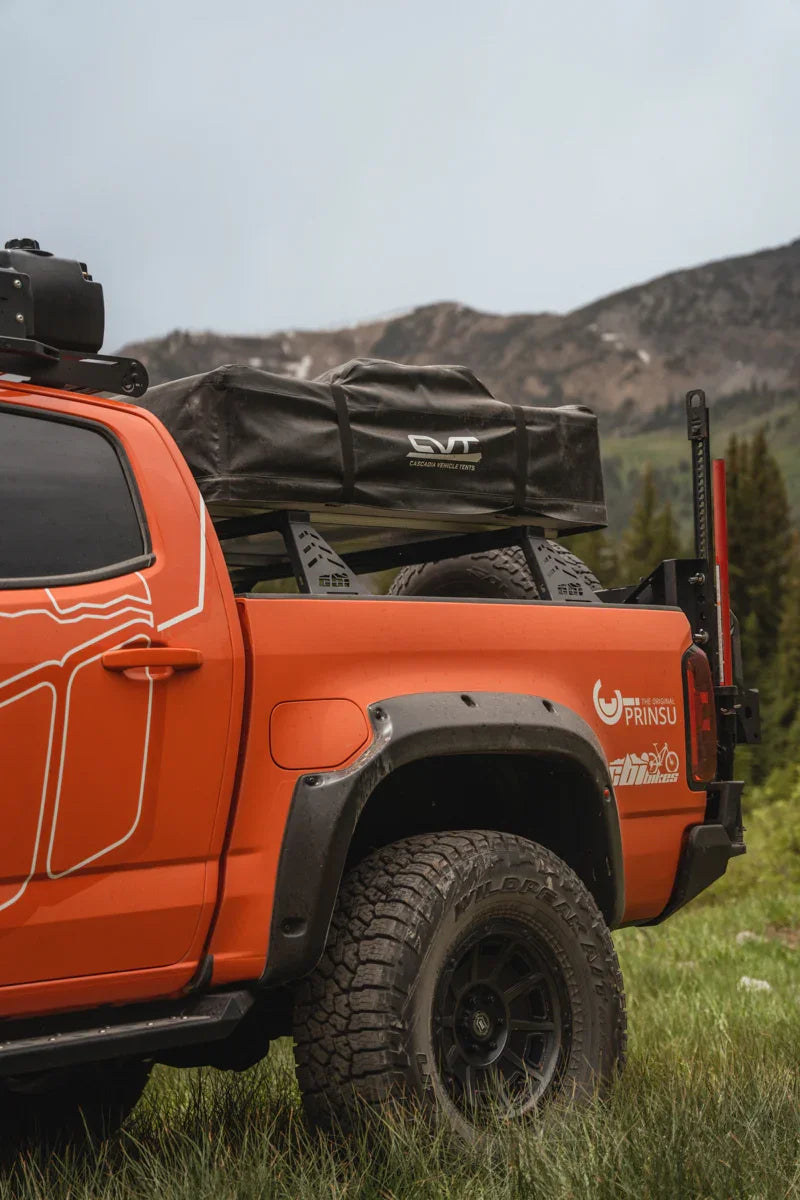 Orange overland truck with bed bars, rooftop tent, off-road tires, and mountain background
