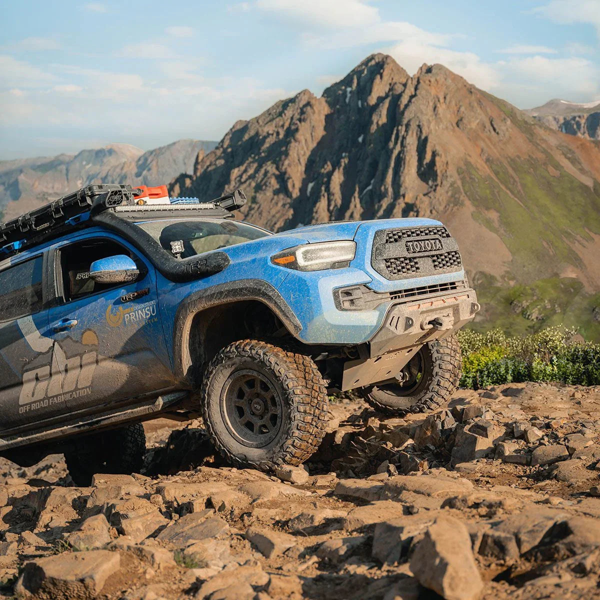 Blue Toyota Tacoma off-road vehicle with roof rack and gear driving on rocky mountain terrain