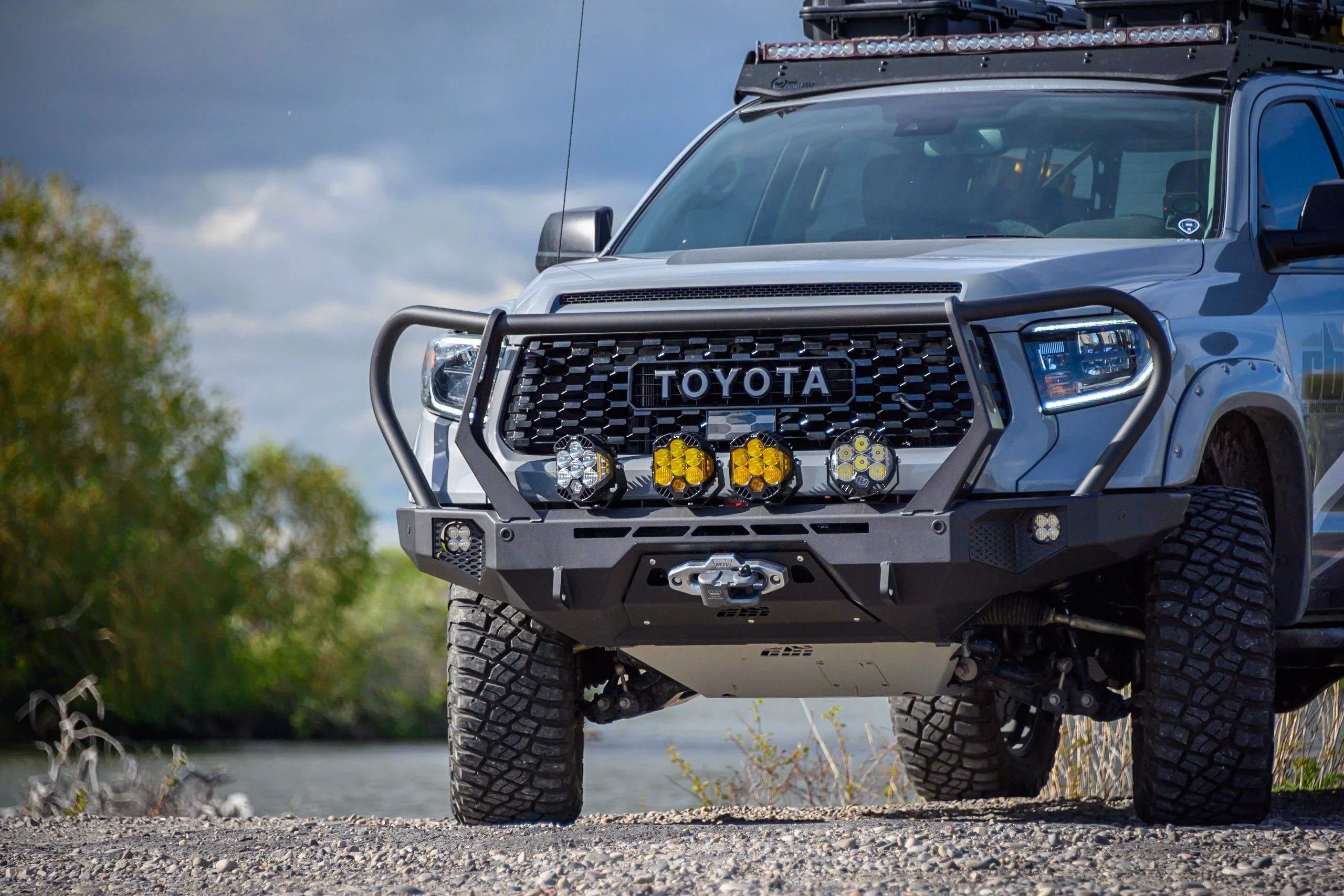 Toyota Tundra with off-road bumper, LED lights, roof rack, and rugged tires by a river