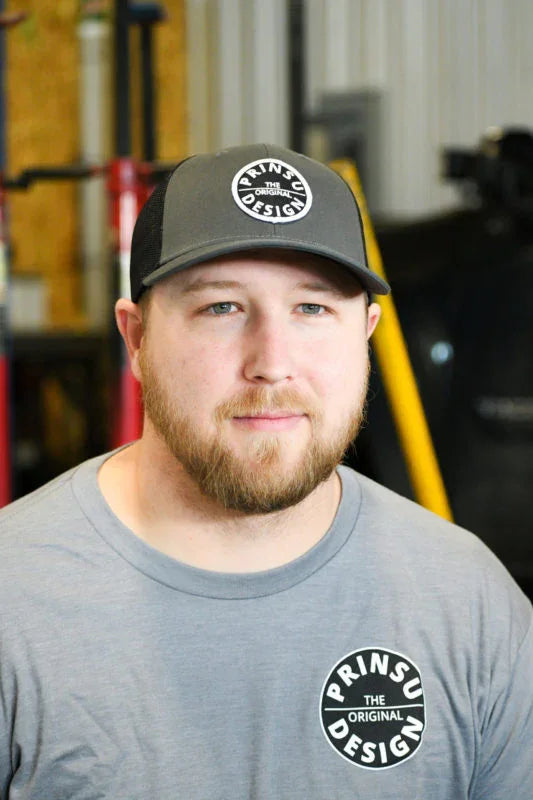 Bearded man in Prinsu Design cap and shirt, indoors, automotive workshop background