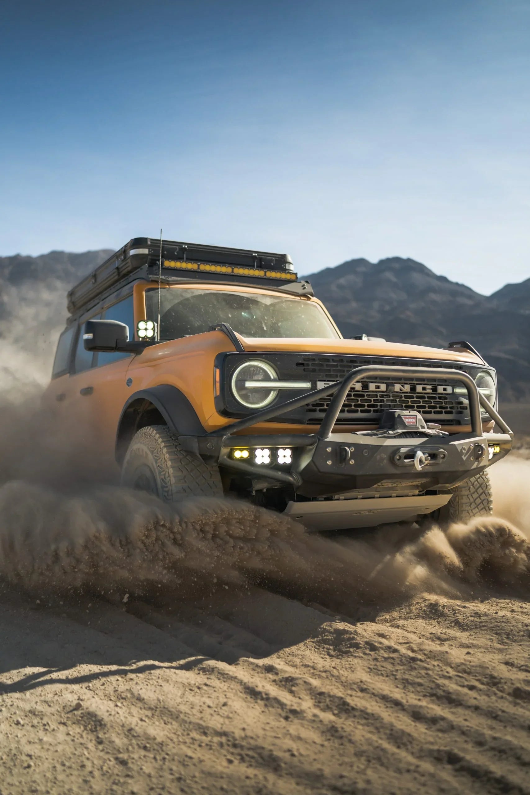 Yellow Ford Bronco with off-road gear driving through desert sand, mountains in background