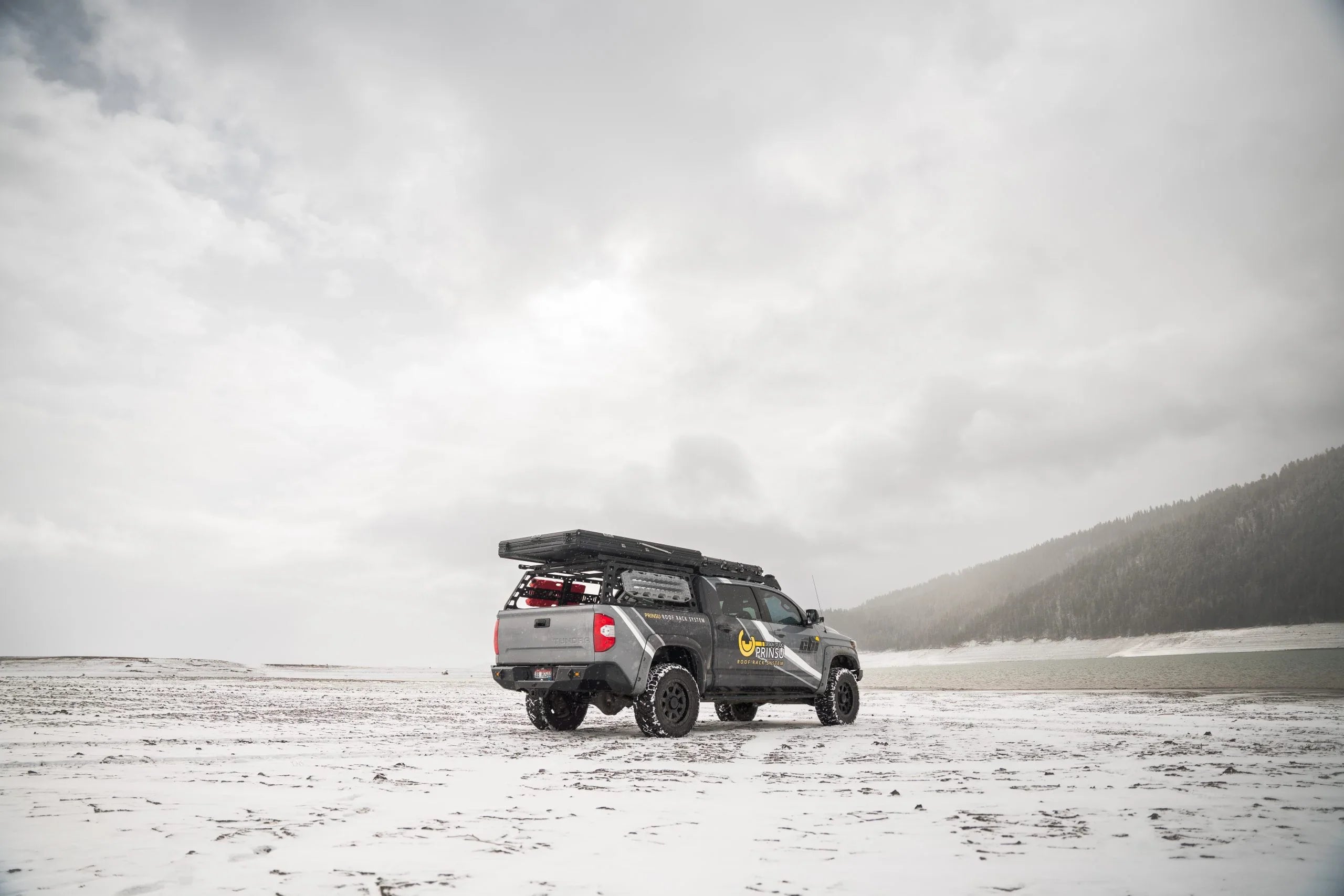 Overland Toyota Tacoma with roof rack and gear parked on snowy landscape by a mountain lake
